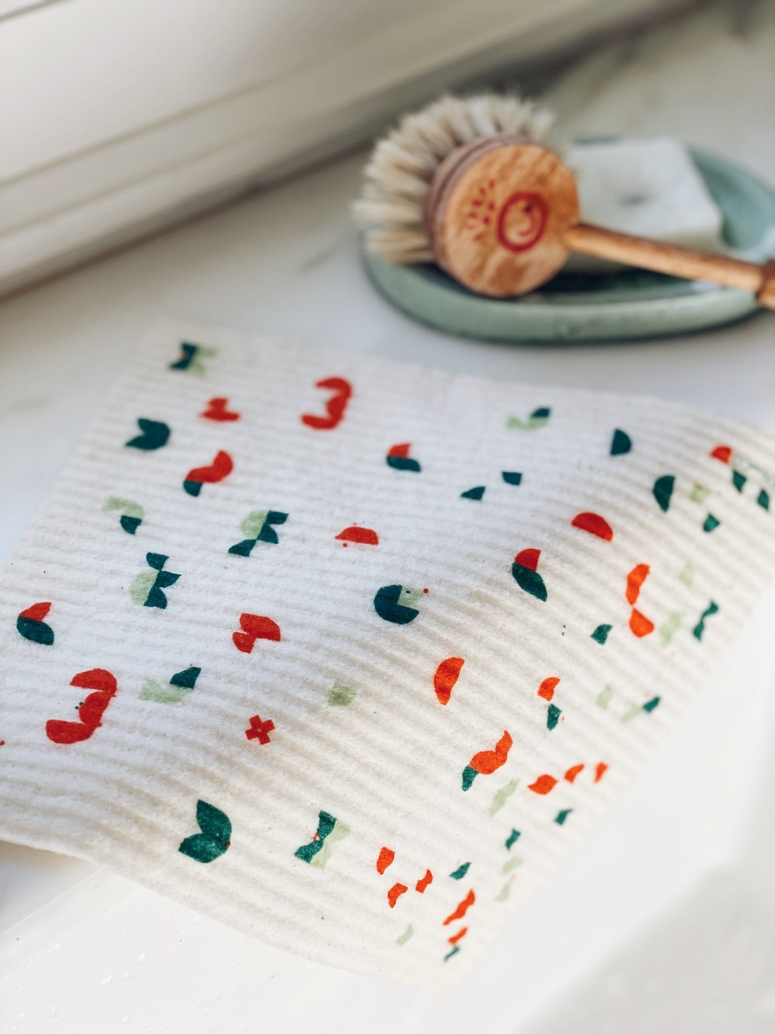 Product image of Geo Animal Sponge Cloth on a white marble surface. The sponge cloth is laying over the edge of a white kitchen sink. There is a soap dish with a dish brush on it in the background out of focus.