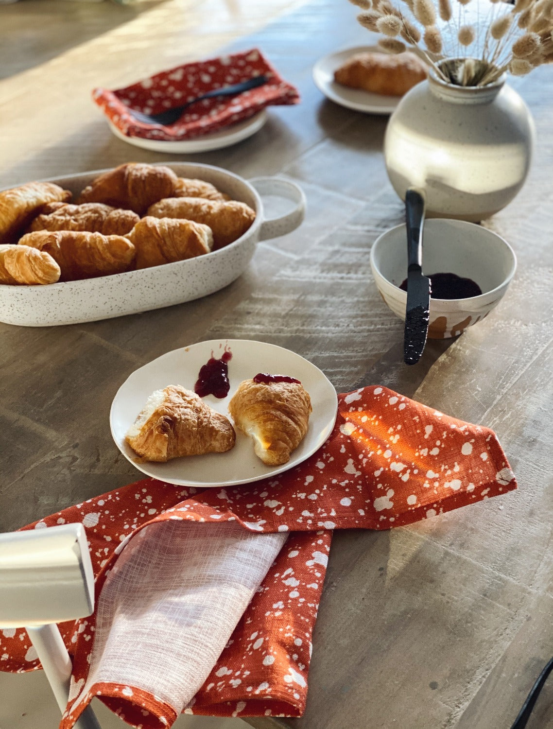 Design image of Splatter Rust napkins shot in natural light. There is a wooden table with a breakfast spread. Clockwise from top left is: a small plate with a folded Splatter Rust napkin on it with a fork, a small plate with a croissant, a vase with some pussy willows, a small bowl with some jam and a knife laying across it, a plate with a croissant cut in half and spoonful of jam on it laying on a Splatter Rust napkin, and an oval white pan with croissants on it. 