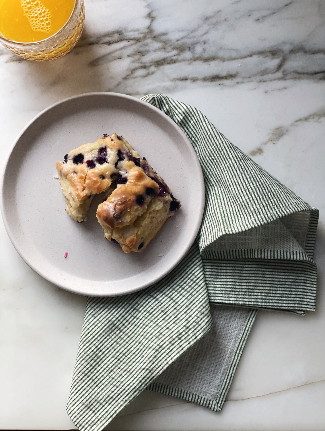 Design image of Stripes Sage napkin on a white marble surface. In the top left is a glass of orange juice. In the middle of the image is a white plate with a blueberry scone on it. The Stripes Sage napkin splayed underneath the plate.