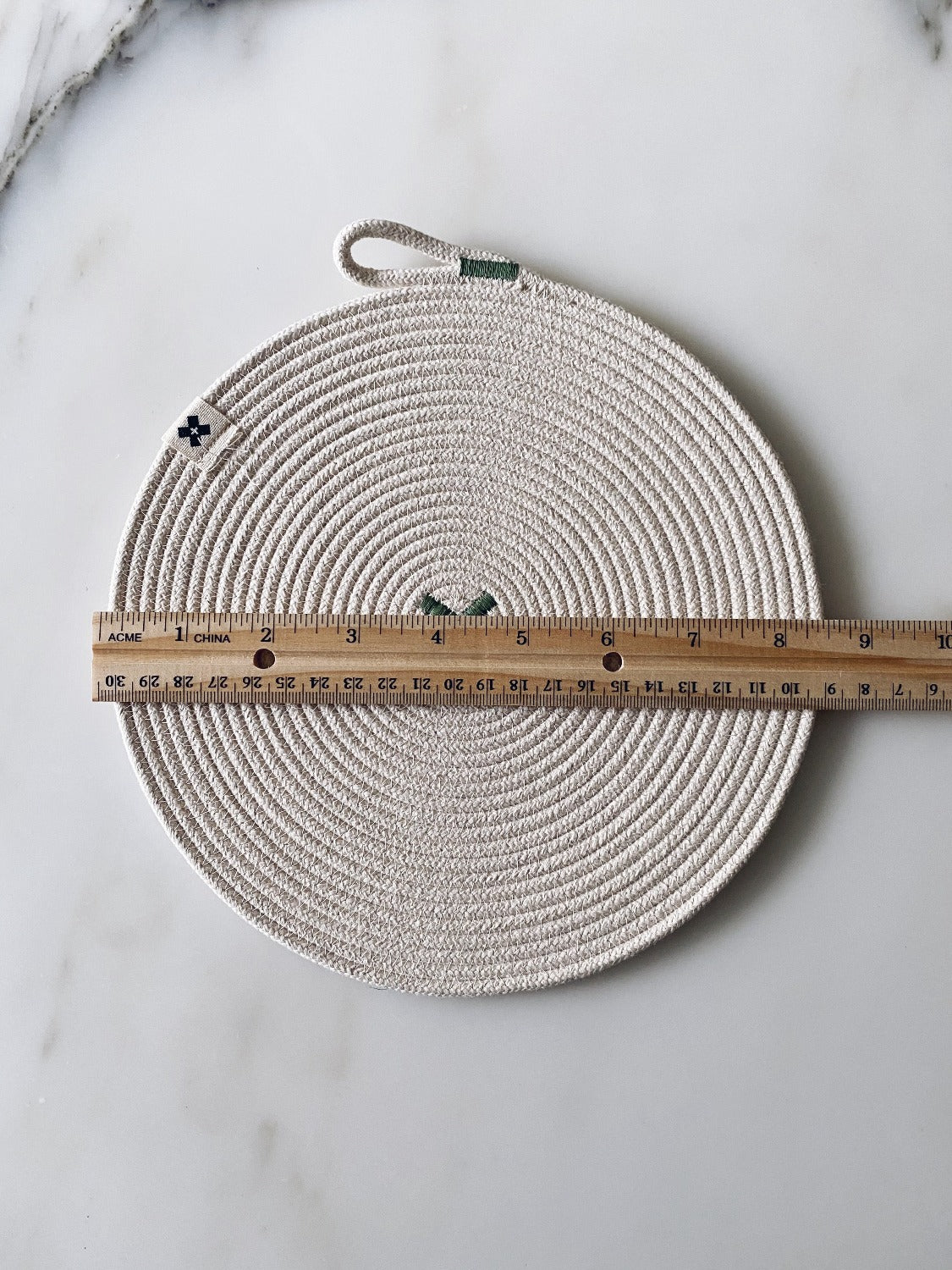 Flat lay image of Large Rope Trivet Sage. The white rope trivet is laying on a white marble surface. There is wooden ruler laying across the trivet. It is about 9 inches in diameter.