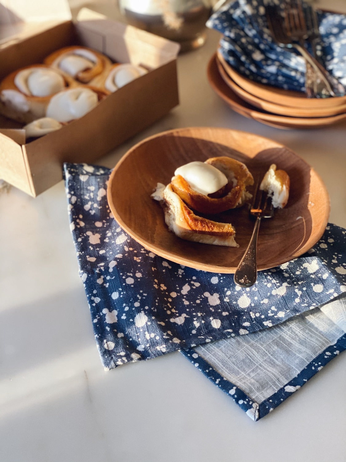 Design image of Splatter Denim napkins shot in natural light. In the top left corner and out of focus is a box of cinnamon buns. Top right corner and out of focus is a stack of wooden plates with a few Splatter Denim napkins laid on top and a pile of forks. Closer to the center of the image and in focus is a Splatter Denim napkin folded once loosely with a wooden plate on top and a cinnamon bun with a fork to it's right and a piece of cinnamon bun on the fork. 