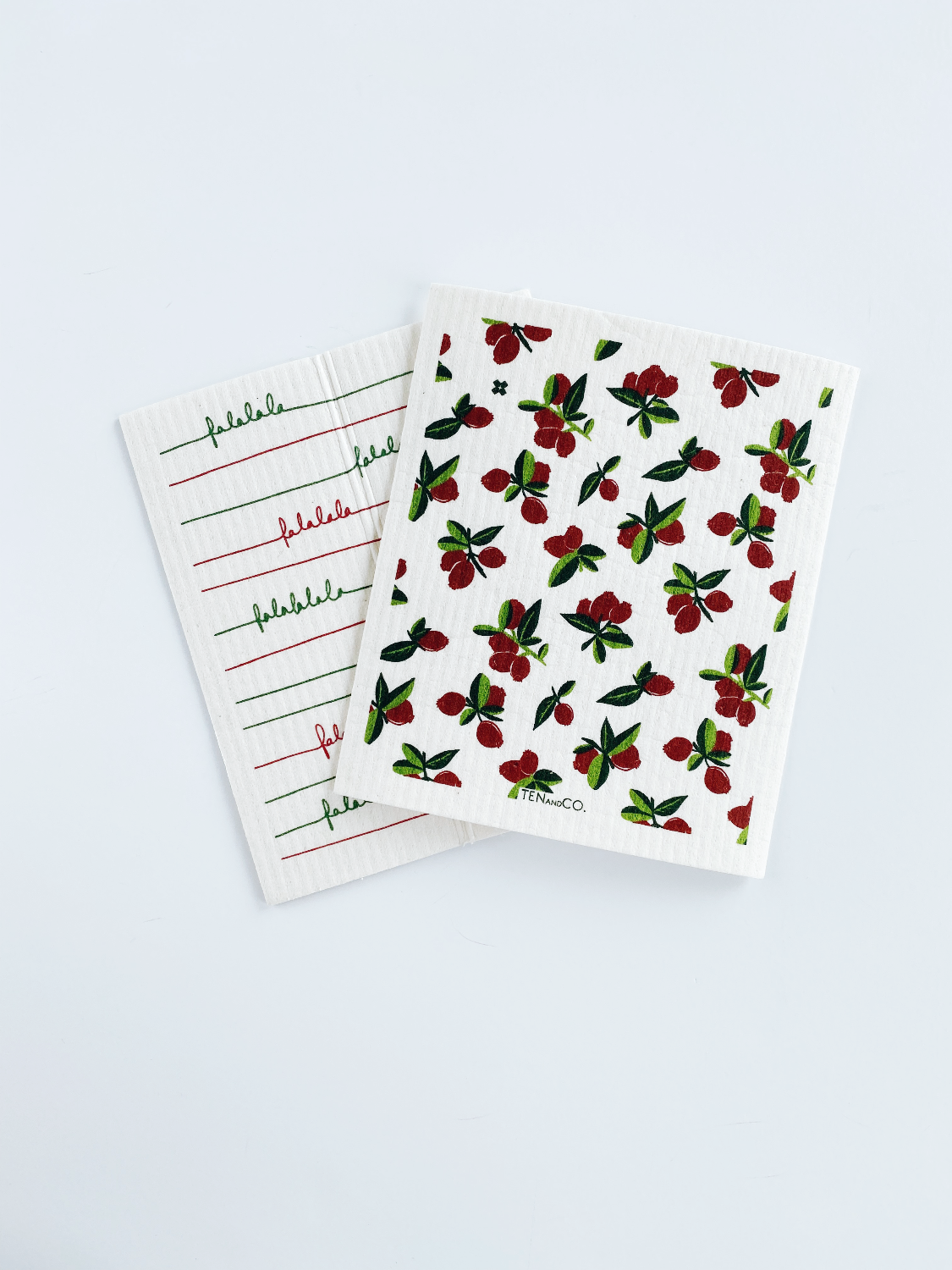 Birds eye view image of Falala Red/Green and Cranberry Sponge Cloths on a white background. Falala cloth is on the left side and slightly slanted and the Cranberry cloth is on the right with it's bottom left corner overlapping the bottom right corner of the Falala Cloth.