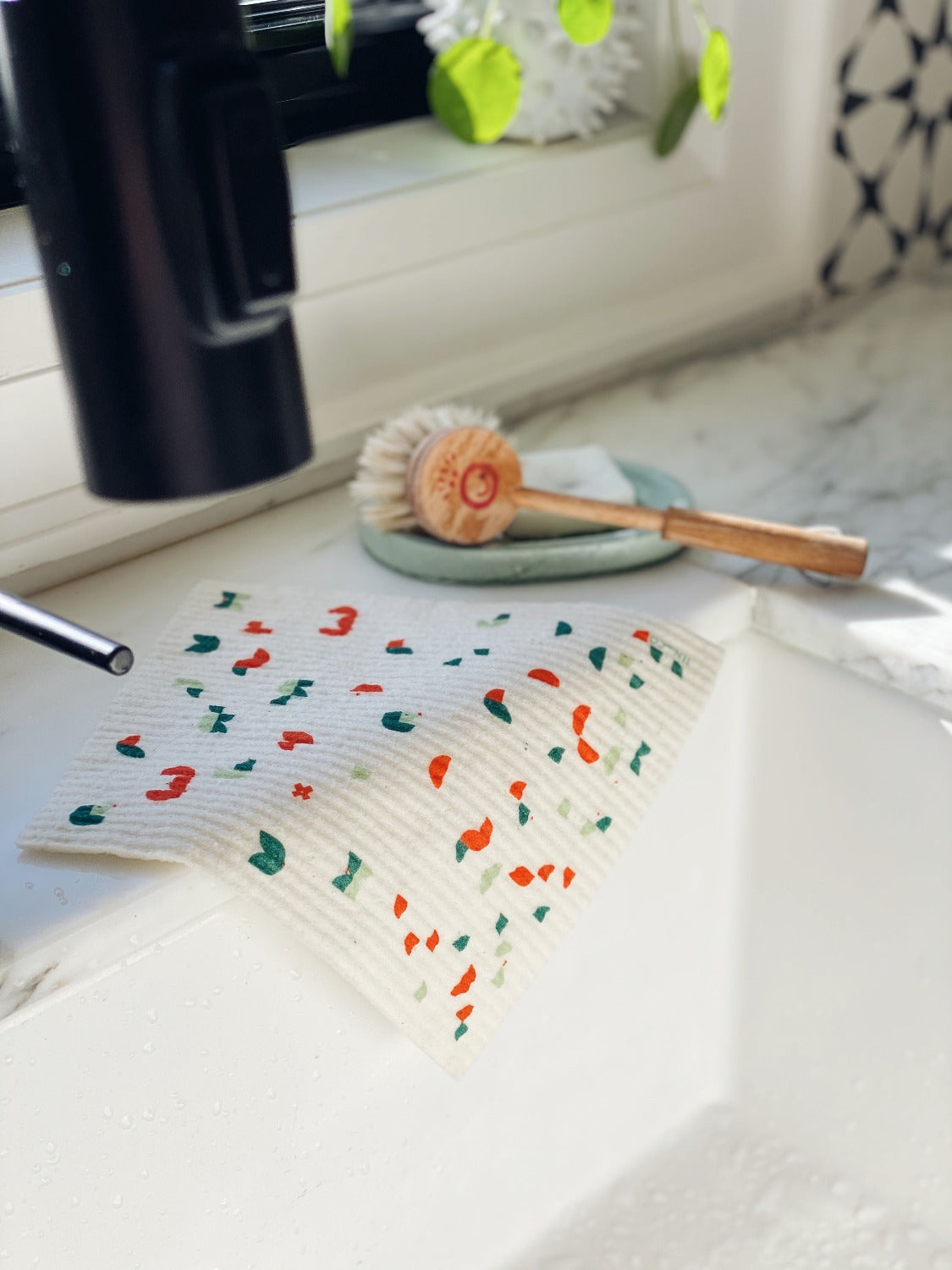 Product image of Geo Animal gift set on a black ground. In this image there is a white kitchen sink. The sponge cloth is laying over the back edge of the sink. There is a soap dish and kids brush beside the cloth on the corner of the sink. You can see the black faucet head in the left corner of the image. There is a window sill towards the back of the image with a small plant on it.