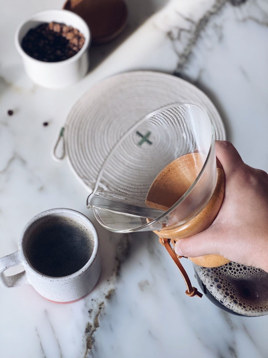 Coffee being poured out of a carafe, the carafe is held high above. In the background is a Sage Trivet, 100% Cotton Canadian Rope Trivet. Towards the top left is a container full of coffee beans. Everything is placed on a marble background, image is viewed through an aerial point of view.