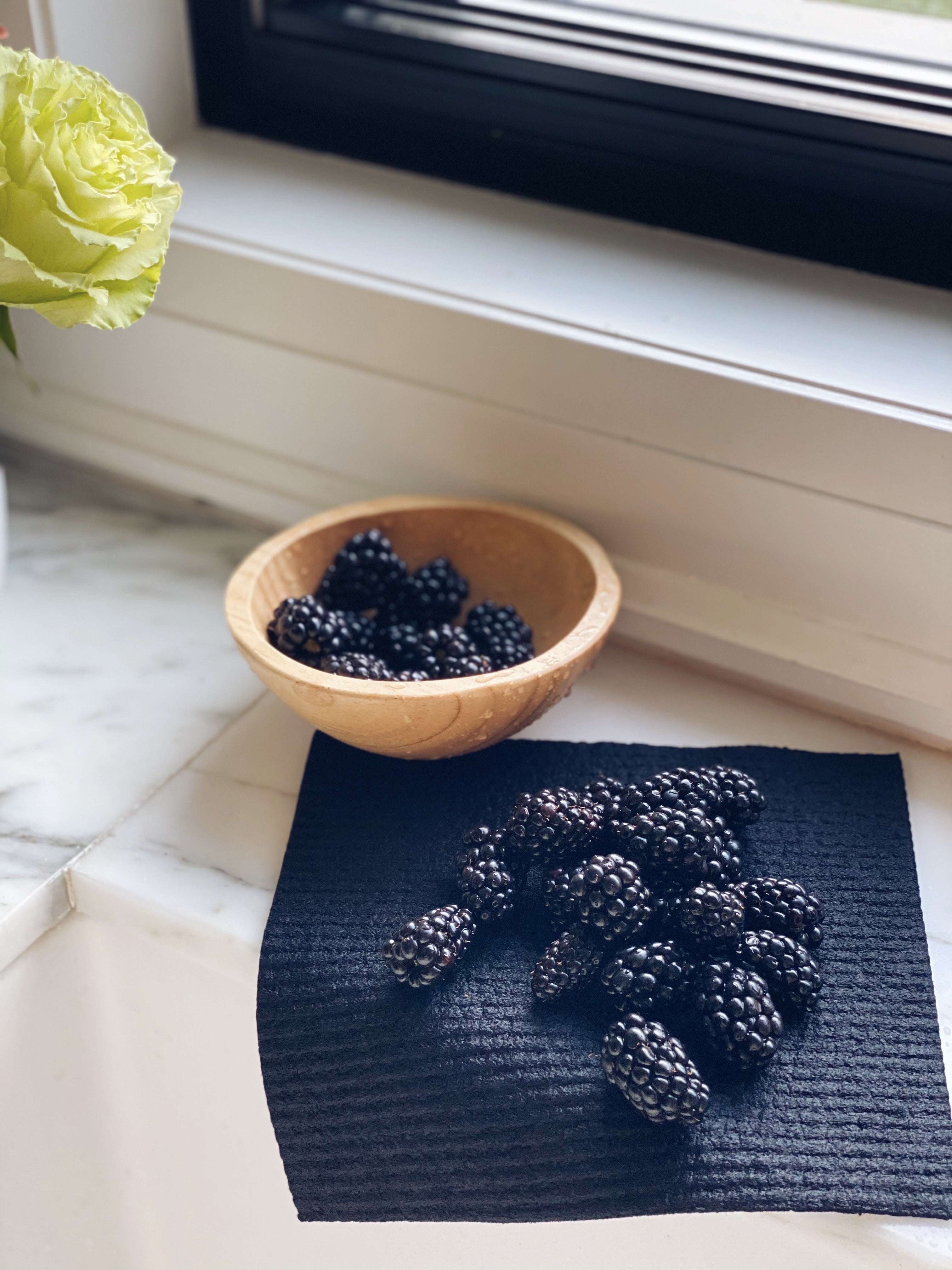 Solid Black Sponge Cloth hangs over the back of the sink. On top of the cloth are freshly washed Blackberry that are drying on the cloth. To the top left of the image is a wooden bowl of blackberries. In the background you can see the light streaming onto the window sill and on the far left is an image of green roses cut off.