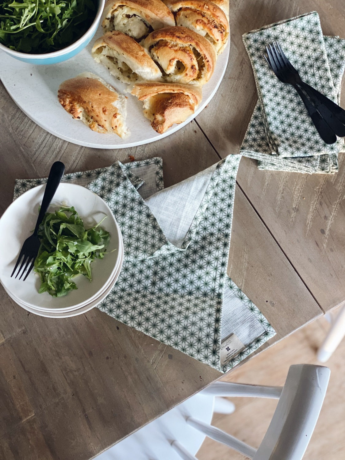 Design image of Starburst Sage napkins on a wooden surface. In the top left of the image there is a plate of food peeking out of the corner. To the left of the image are three small, white stacked plates with a metal fork on it and some salad. The Starburst Sage napkin is splayed underneath. To the right of the image is 3 Starburst Sage napkins stacked with 3 metal forks laying vertical. There is a white chair tucked underneath the table as well.