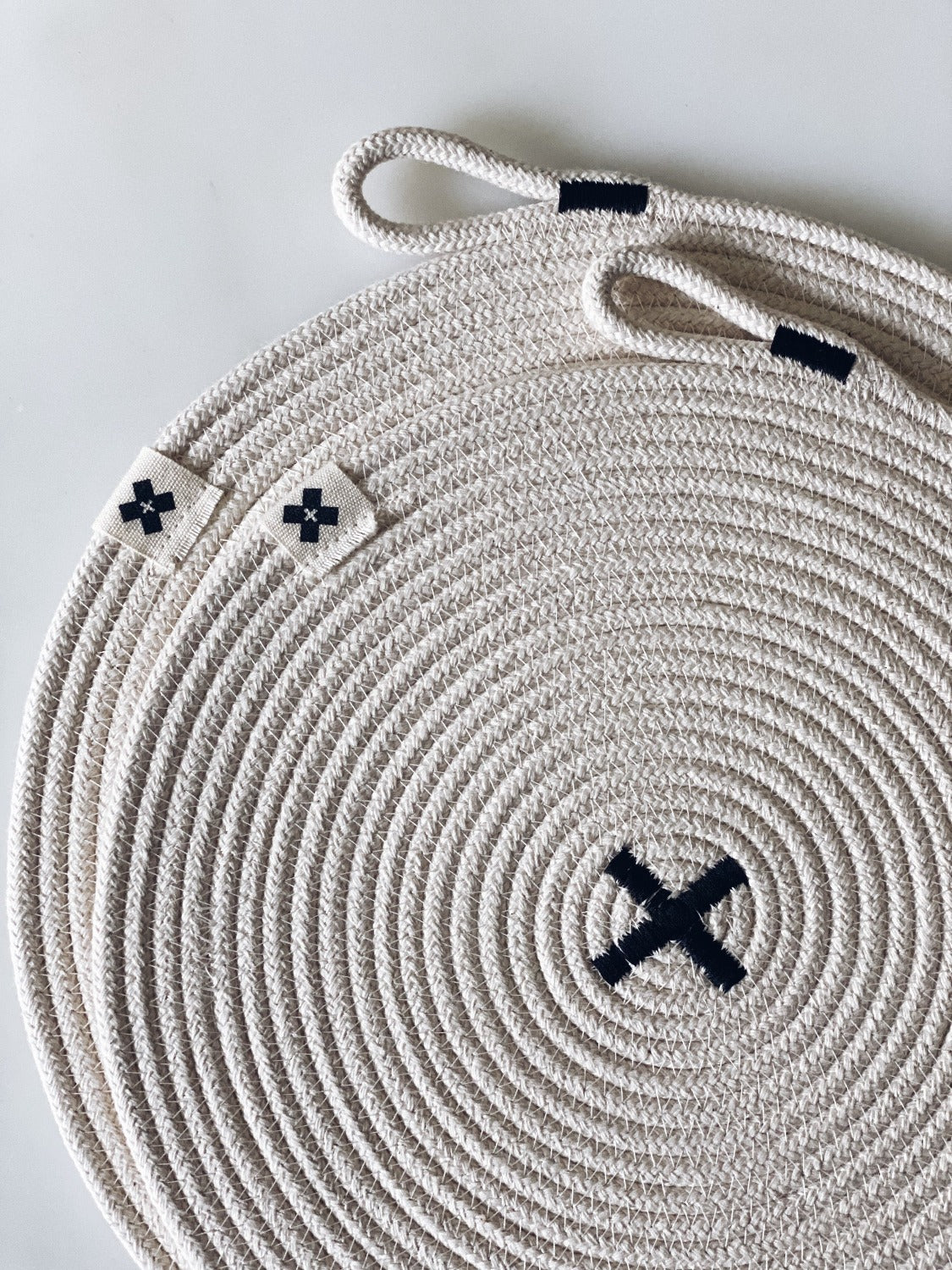 Close up product image of Large Rope Trivet | Black on a white background. In this image there are two different sized trivets on top of each other. You can see the X in the middle of the top one and the tiny Ten and Co. tag attached to each.