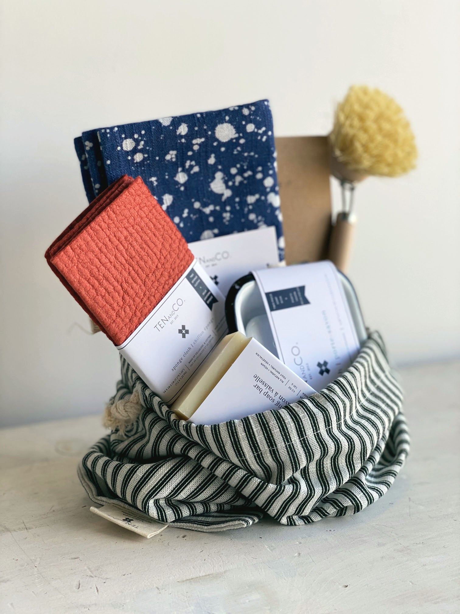 Angled photo of Gift Bag with Ten and Co items inside. Photo on a white surface against a white wall. Deep green and white vertical striped gift bag is propped open with the following items inside. Left to right; Rust solid 2pk, Splatter Denim Napkins, Beeswax Wraps, Scrub Brush. In front of these items in the bag, is a Dishwashing Soap Bar and an Enamel Soap Dish.