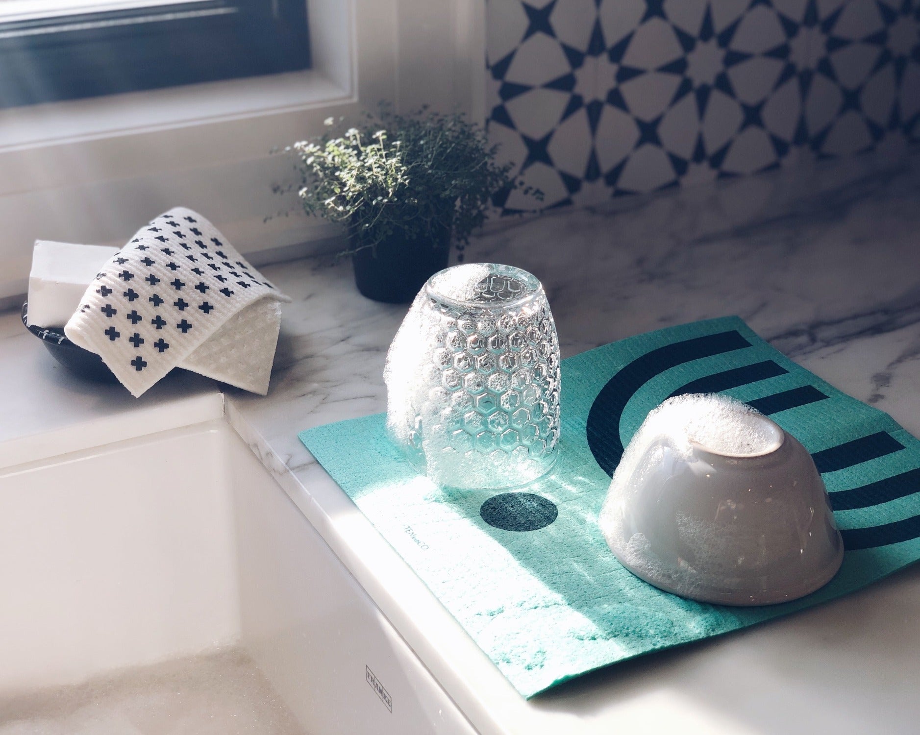 Product image of LARGE Arc Green Sponge Cloth Mat. There is a marble counter beside a white kitchen sink. On the counter is a LARGE Arc Green sponge cloth mat with a glass and a bowl upside down, drying. There is a soap dish with a Tiny X sponge cloth on it. Beside the soap dish is a small green plant. Above the sink is a window with sun shining through on the sponge cloth mat. 