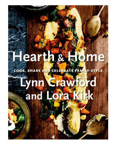 Flat lay image of Hearth and Home book by Lynn Crawford and Lora Kirk. The background picture is a dinner spread on a cutting board. There are two large spoons and a small plate with food on it. “Hearth & Home: Cook, Share and Celebrate Family-Style" is in bold white font across the middle of the book. The authors names are below the title.