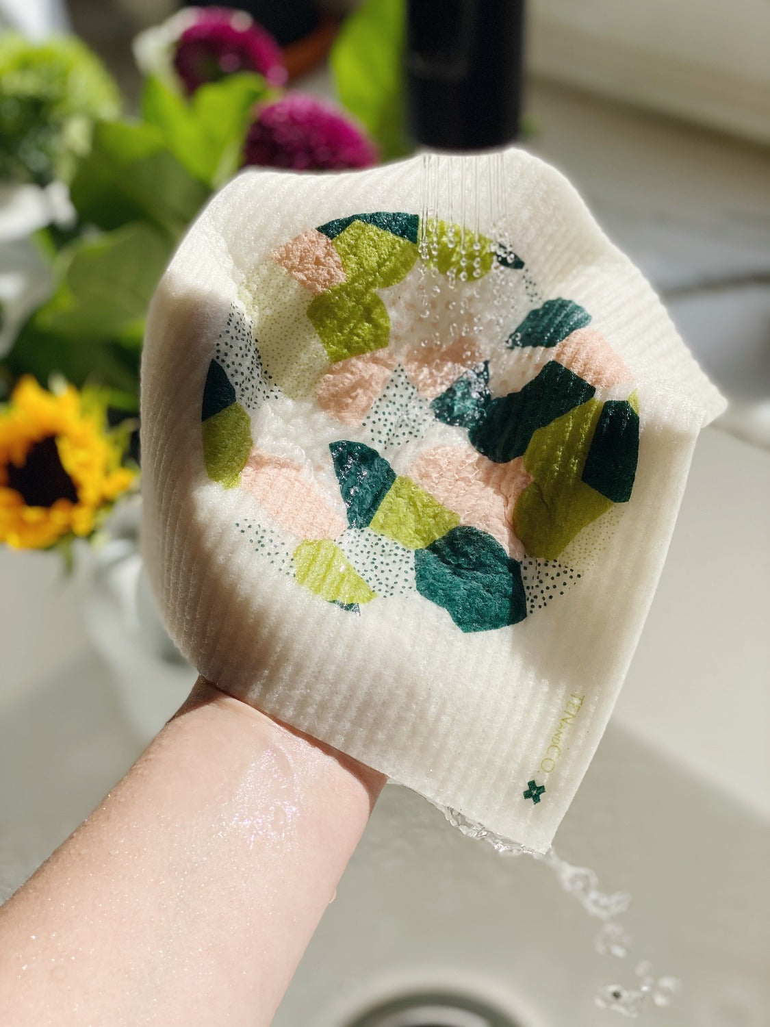 Product image of Full Circle Sponge cloth. There is a hand coming out of the bottom of the image over a kitchen sick. There is sunlight shining on the wet, drippping sponge cloth. There is a bouquet of flowers in the background in the top left.