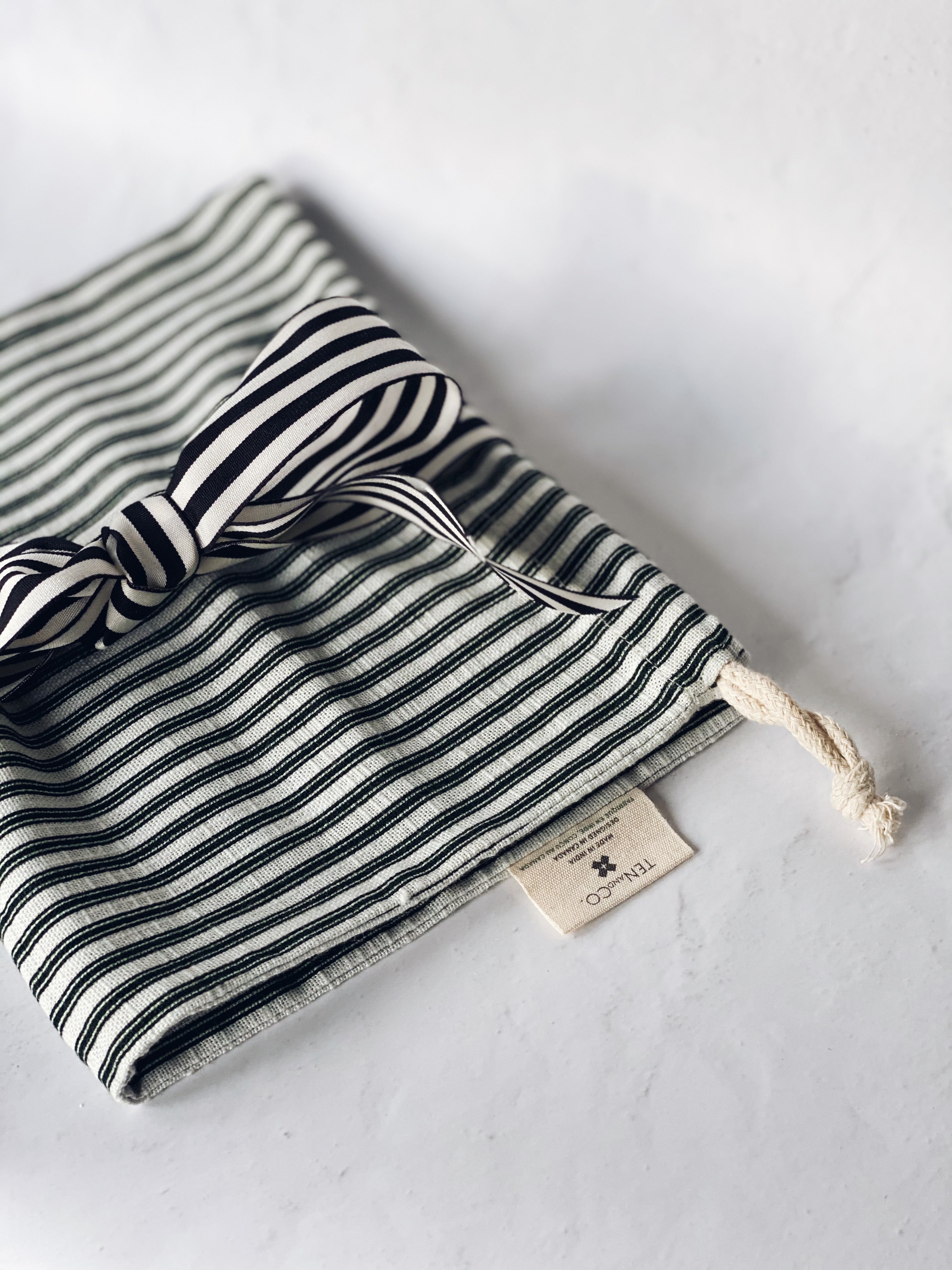 Angled photo of Gift Bag laying flat, folded in half, on a marbled surface. The deep green and white striped Gift Bag is loosely tied around the center with a black and white striped ribbon, formed into a bow. The Ten and Co tag and drawstring from the bag are showcased closer to the bottom of the image. There is some shadowing around the bag in this image.
