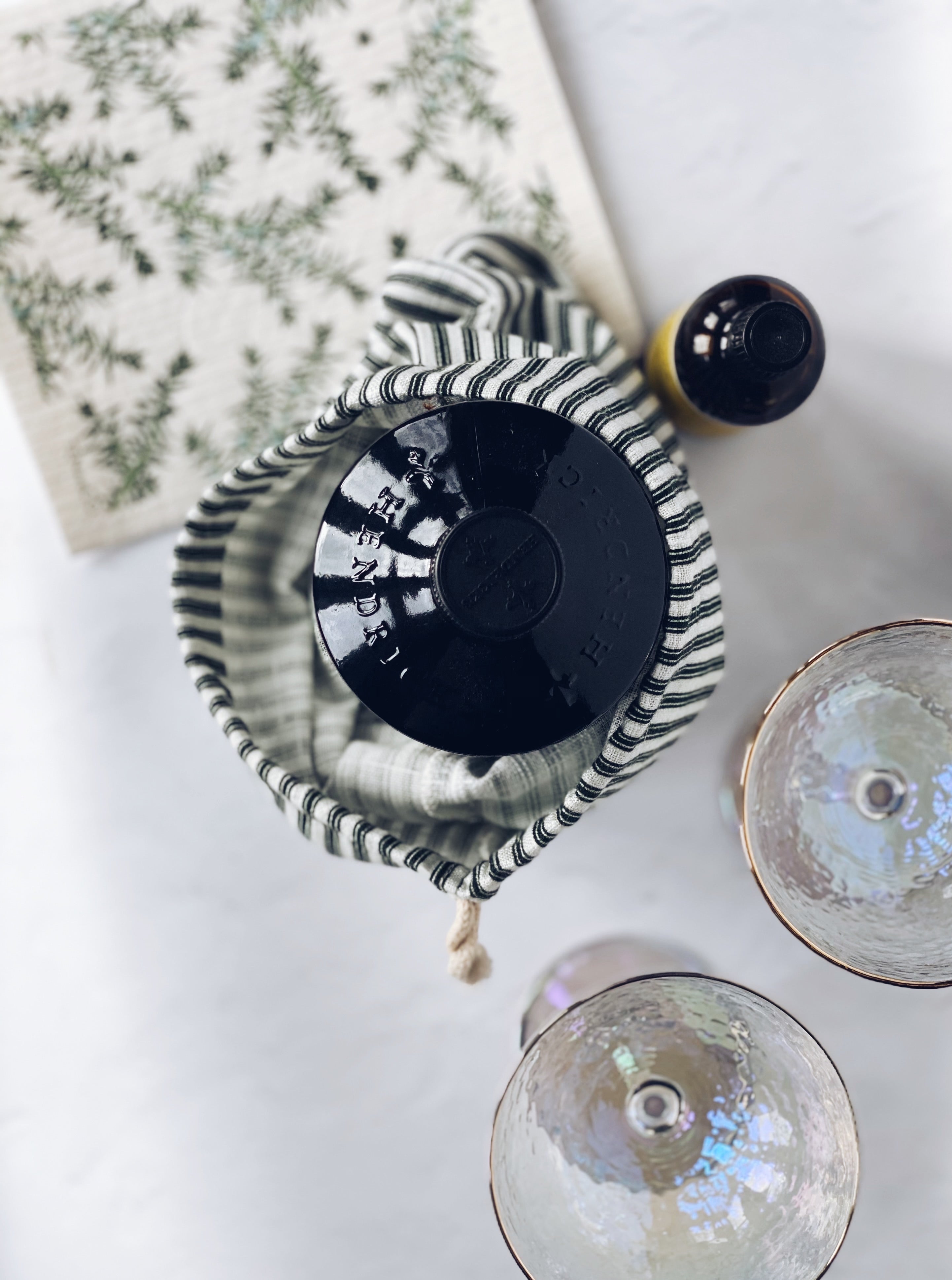 Birds-eye view photo of a french press inside of the Gift Bag. Center and focus of the photo is the top of a french press seen inside the gift bag. In the top left corner out of focus is a Juniper Greens on White Sponge Cloth. To the top right of the french press is a small glass jar with a yellow label. In the bottom right of the image is 2 stemmed, textured empty glasses with a gold rim. All items are on a white surface with some natural light coming from the left side.