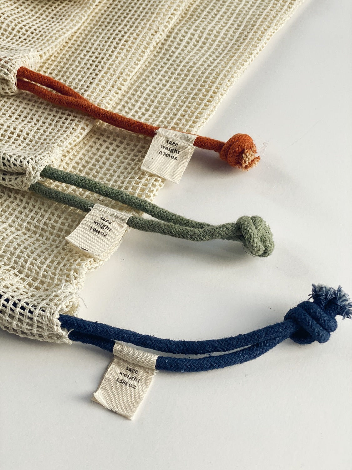 Flay lay image of Produce Bags on a white background. This is a close up image of the string colours with the small Ten and Co. Tags. Each drawstring is knotted at the end. The small is rust, the medium is green and the large is blue. 