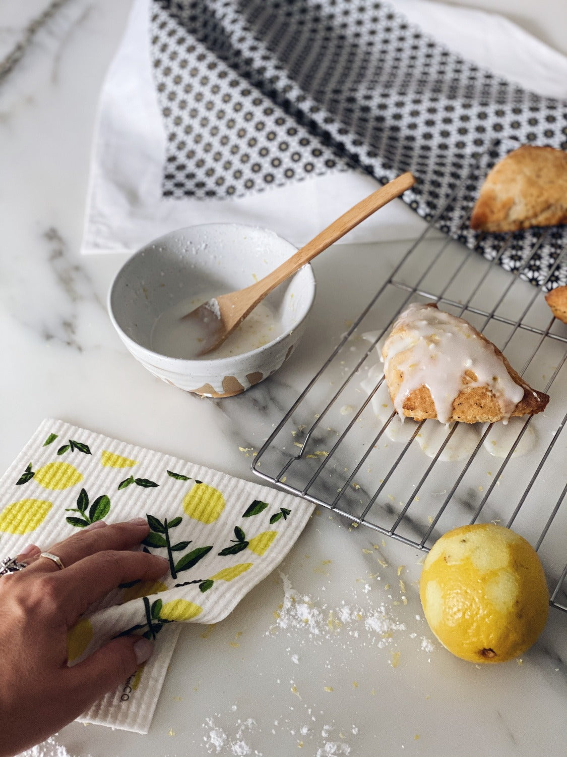 Product image of Citrus Lemon Gift set on a white marble surface. In this image there is a hand holding the sponge cloth wiping up some flour. There is a Starburst Blue tea towel at the top with a scone on it. Below that is a small bowl with icing in it and a wooden spoon. Beside the bowl is a wire rack that has a scone on it with icing dripping from it. There is a lemon that has been zested beside the wire rack. 