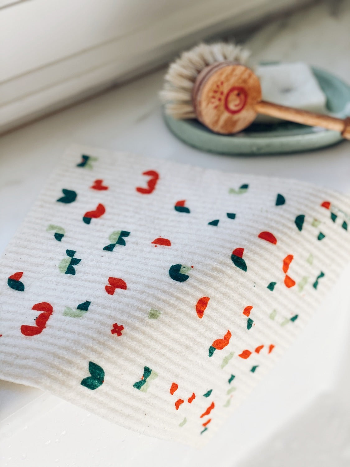 Product image of Geo Animal Sponge Cloth on a white marble surface. The sponge cloth is laying over the edge of a white kitchen sink. There is a soap dish with a dish brush on it in the background out of focus. 