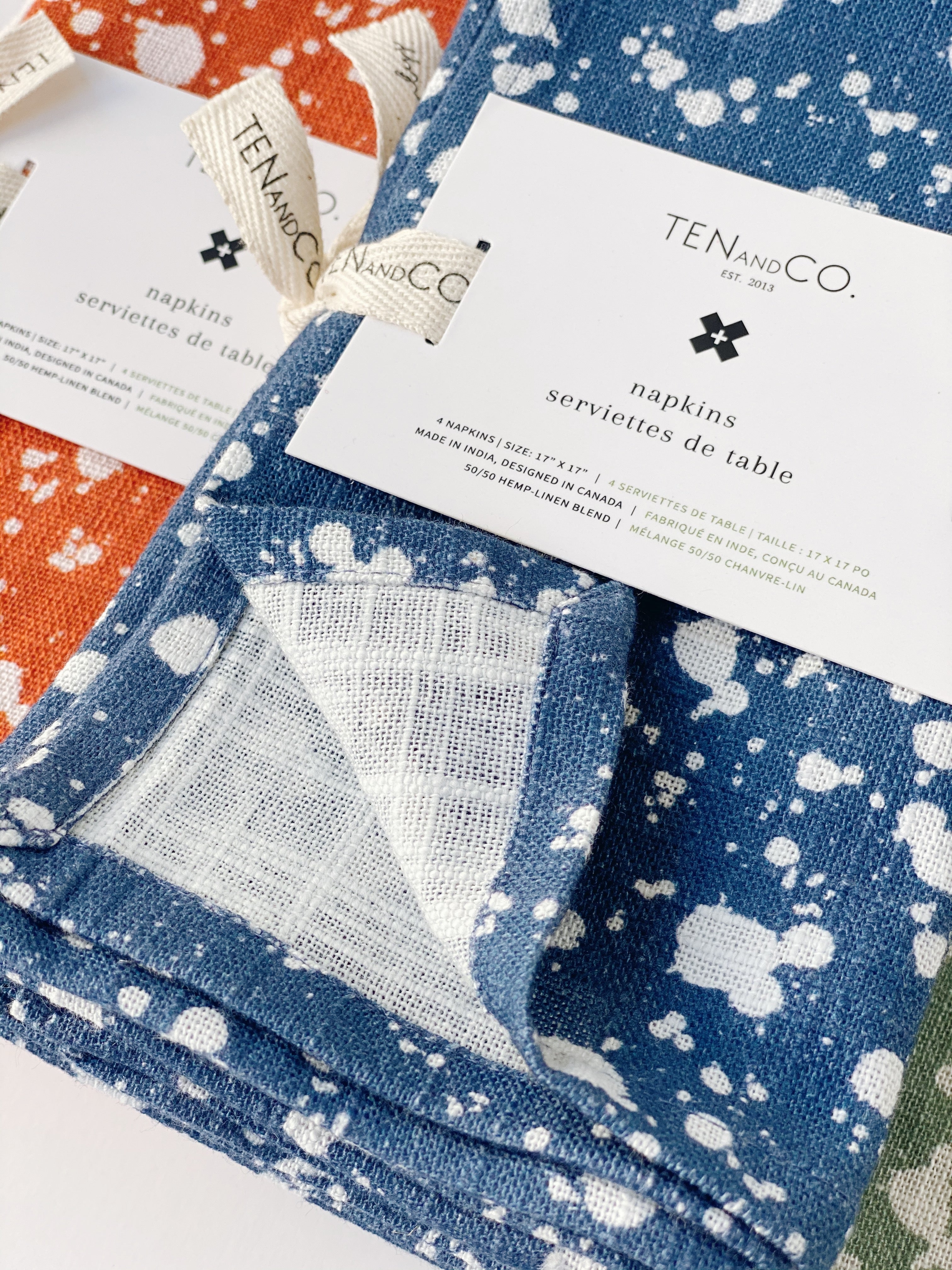 Focus image on the Splatter Denim set of napkins. Splatter Rust napkins are shown slightly in behind the Splatter Denim napkins, and in the top left corner. The focus of the image is the Splatter Denim napkins, tied with a Ten and Co printed description label on the front.