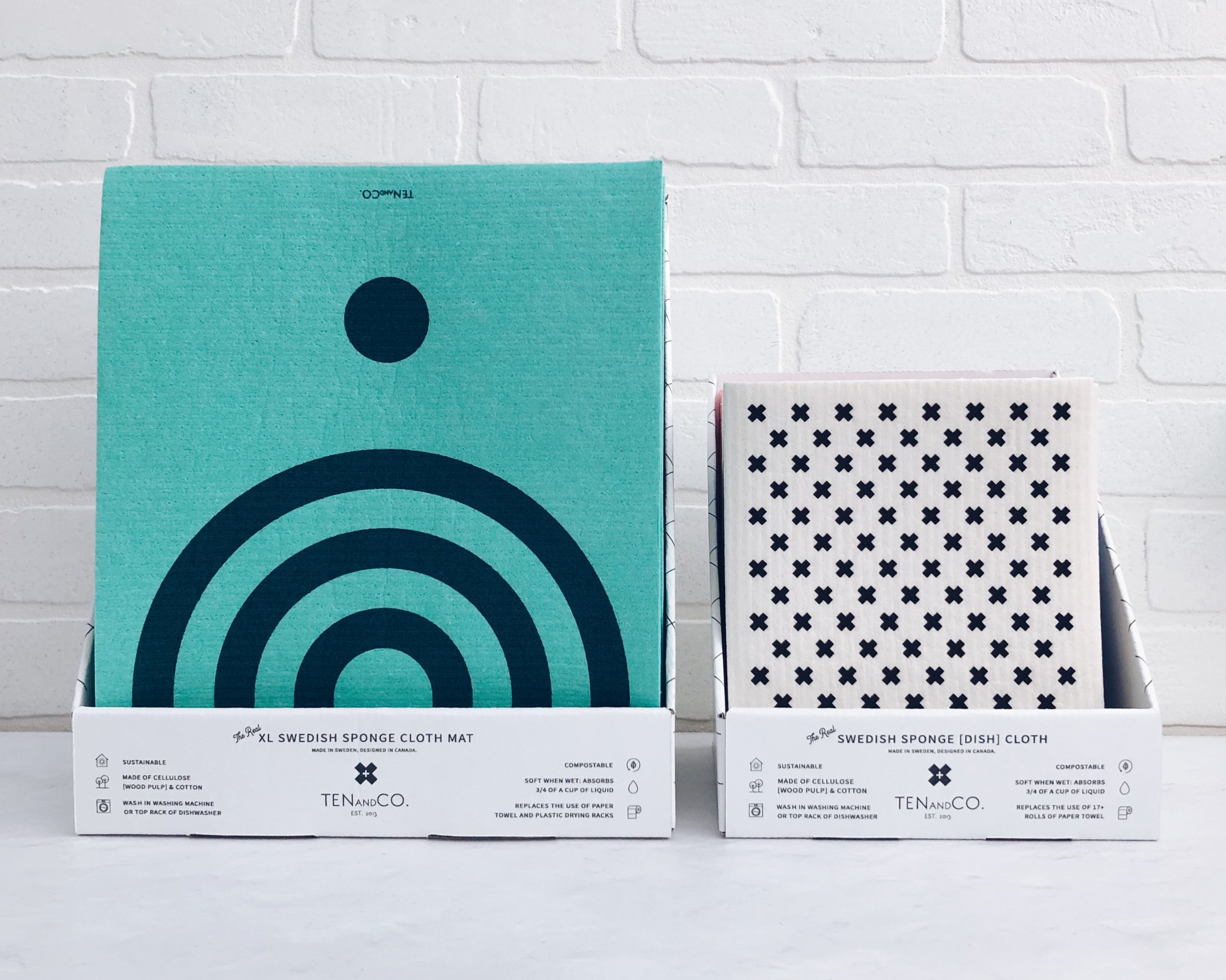 Straight on image of the Cardboard Display Box for Drying Mats and Cardboard Display Box for Sponge Cloths. Both boxes are on a white surface with white brick background. The Drying Mats display is on the left side, whereas the Sponge Cloth display is on the right side. The Drying Mat at the front of the display box is Arc Navy on Blue and the Sponge Cloth at the front of the display box is Tiny X Black/White. 