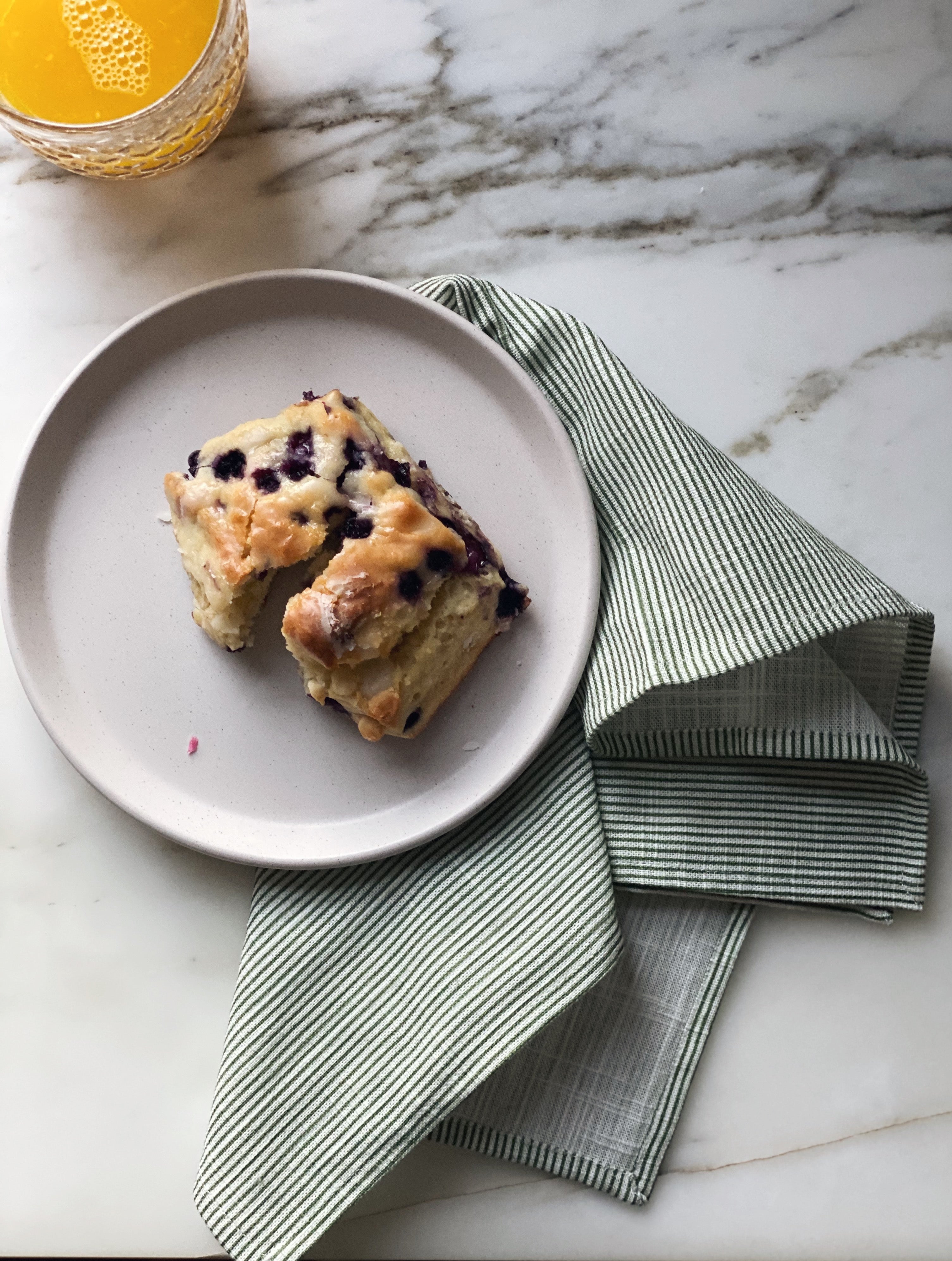 Design image of Stripes Sage napkin on a white marble surface. In the top left is a glass of orange juice. In the middle of the image is a white plate with a blueberry scone on it. The Stripes Sage napkin is splayed underneath the plate.