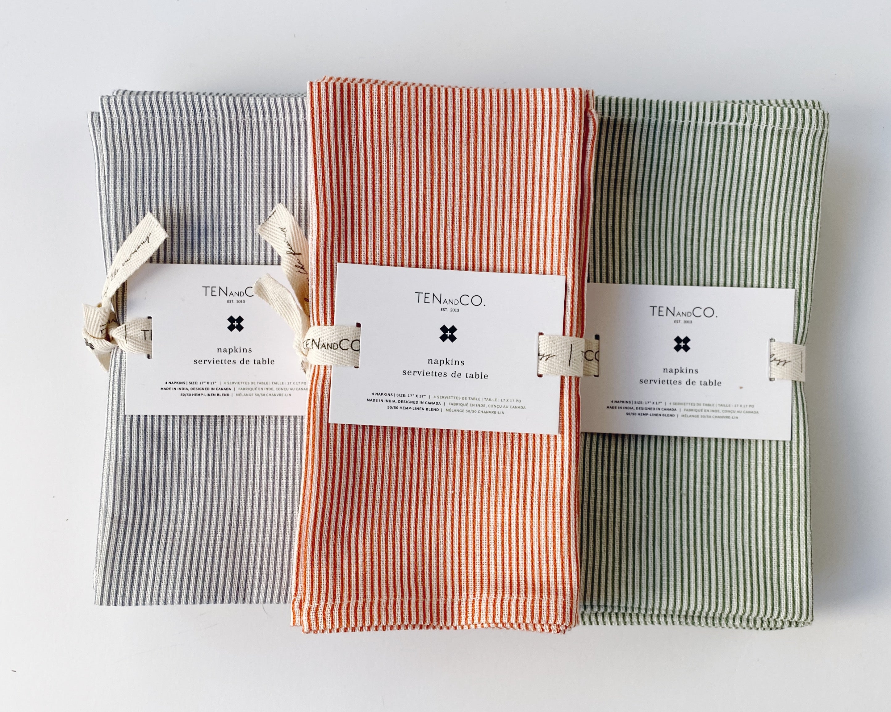 Flat lay image of 3 different sets of napkins displayed vertically. From left to right; Stripes Denim, Stripes Rust, Stripes Sage. All napkins are tied with the Ten and Co printed description label on top.
