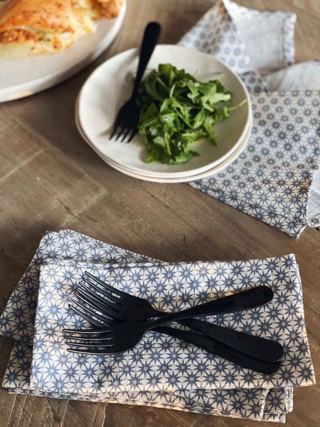Design image of Starburst Denim napkins on a wooden surface. In the top left of the image there is a plate of food peeking out of the corner. There are two small white stacked plates with a metal fork on it and some salad. The Starburst Denim napkin is splayed underneath. At the bottom of the image is 3 Starburst napkins stacked with 3 metal forks laying vertical. 