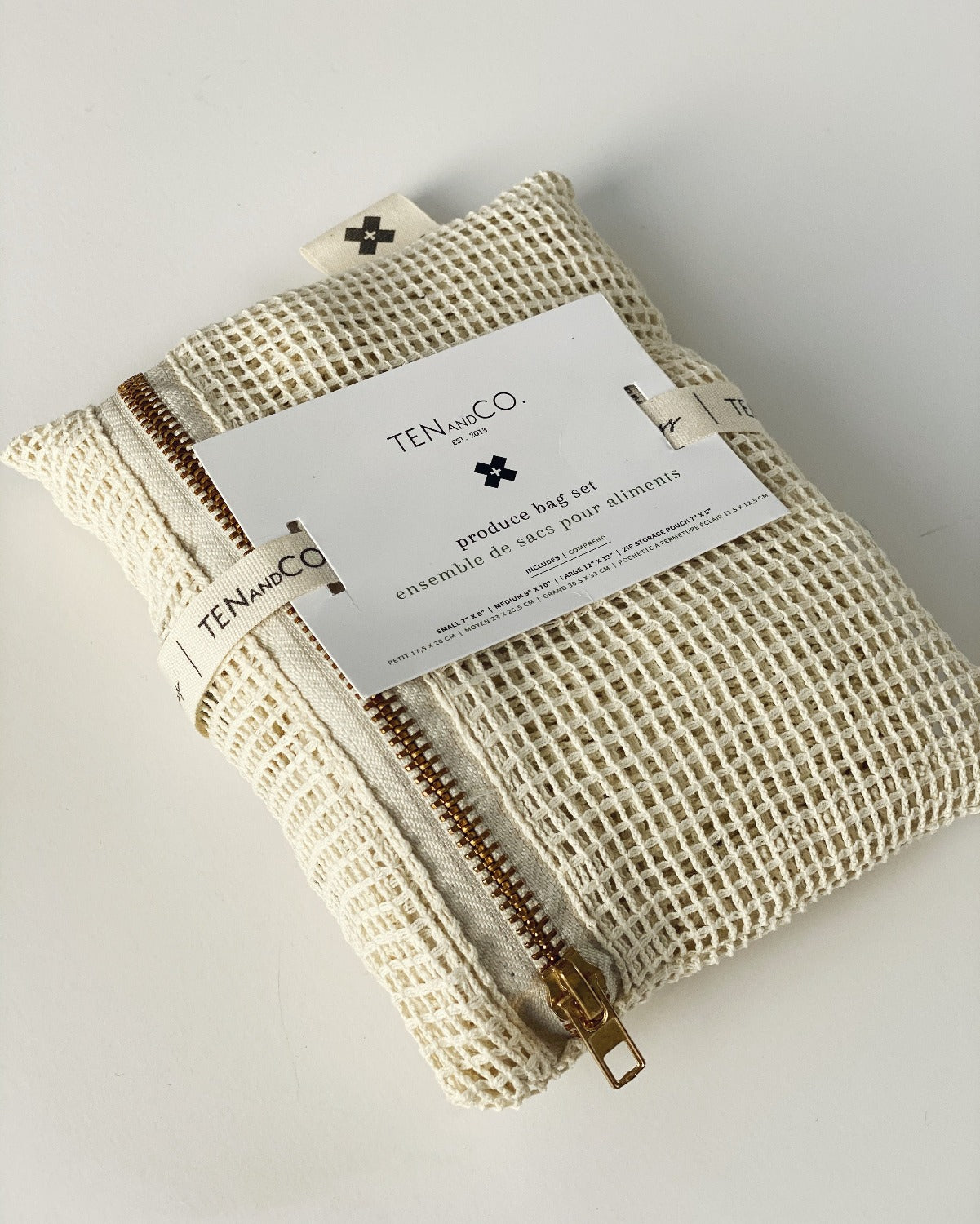 Flay lay image of Produce Bags on a white background. This is the close up image of the cotton mesh pouch with the produce bags inside. The pouch is zippered and has a paper tag attached by cotton ribbon. The tag has the Ten and Co. Logo on it with descriptive text in black font. 