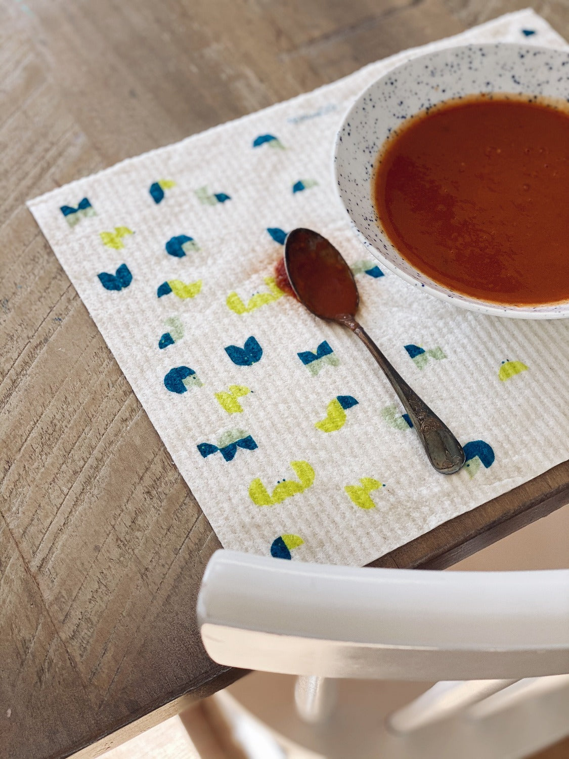 Product image of LARGE Geo Animal Sponge Cloth Mat. In this image there is a wooden table with a Large Geo Animal Sponge cloth being used as a placemat. There is a bowl of red soup with a metal spoon beside it. There is a white chair tucked in to the table. 