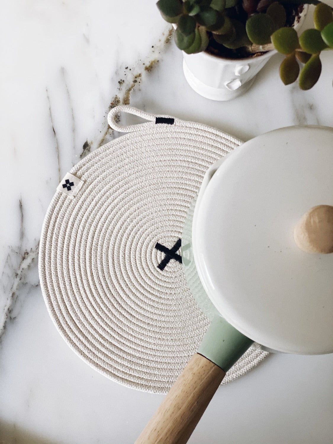 Aerial product image of Large Rope Trivet | Black. On a white marble surface there is a large trivet with a white pot on the edge. The pot has a lid and a wood handle. Above the trivet is a small white planter with a green plant.
