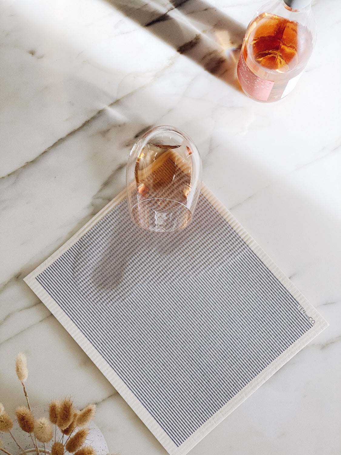 Product image of LARGE Stripe Denim Sponge Cloth mat. The Stripe Denim sponge cloth mat is on a white marble surface. There is a glass with wine that has spilt on the sponge cloth mat. There is a bottle of wine beside the mat. There are pussy willows in the bottom left corner. 