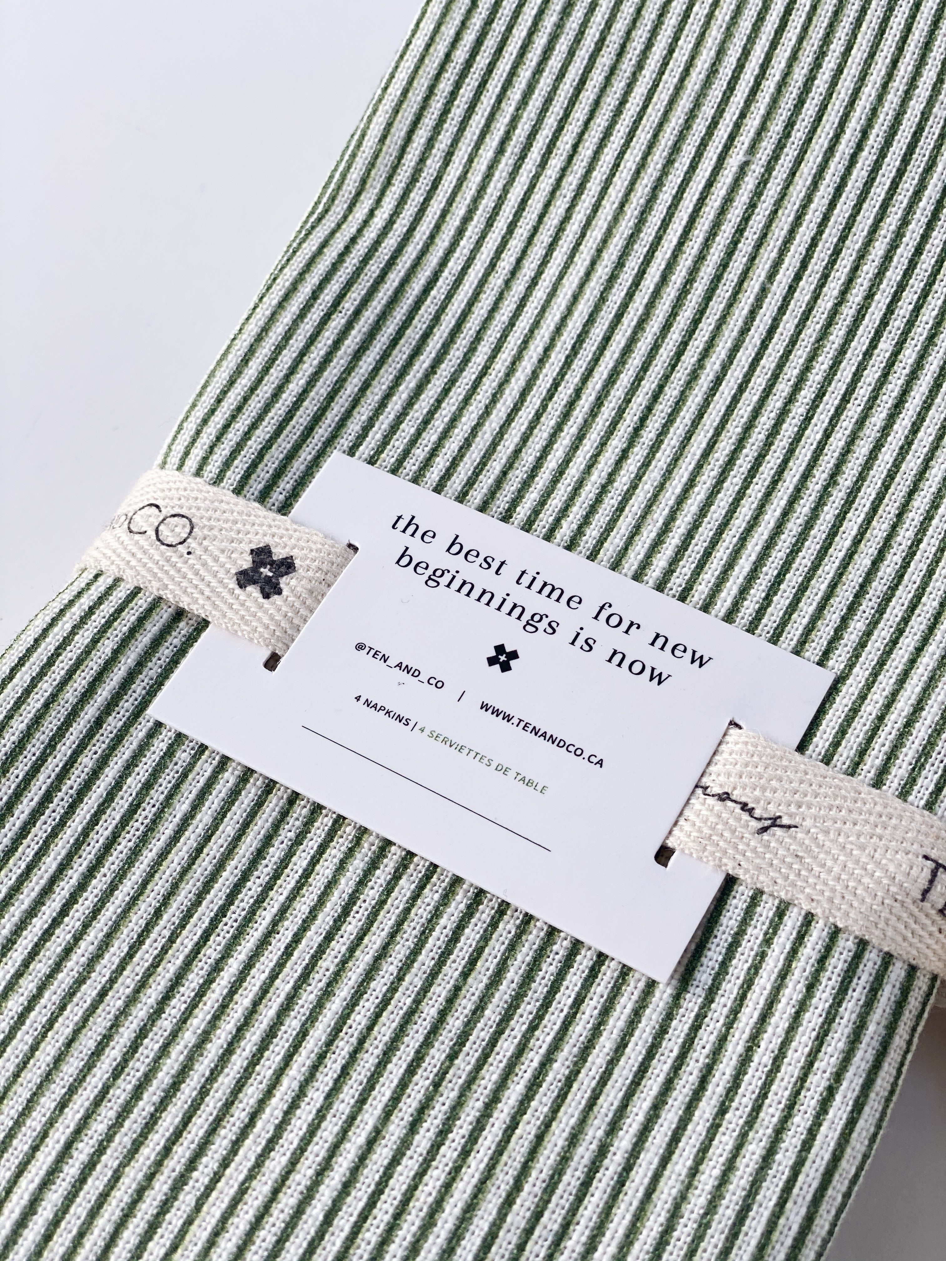 Focus image of Stripes Sage napkins on an angle. Tied together with Ten and Co twill tape and a recyclable paper tag laying on top. The tag reads “The best time for new beginnings is now” in black font with descriptive information underneath.