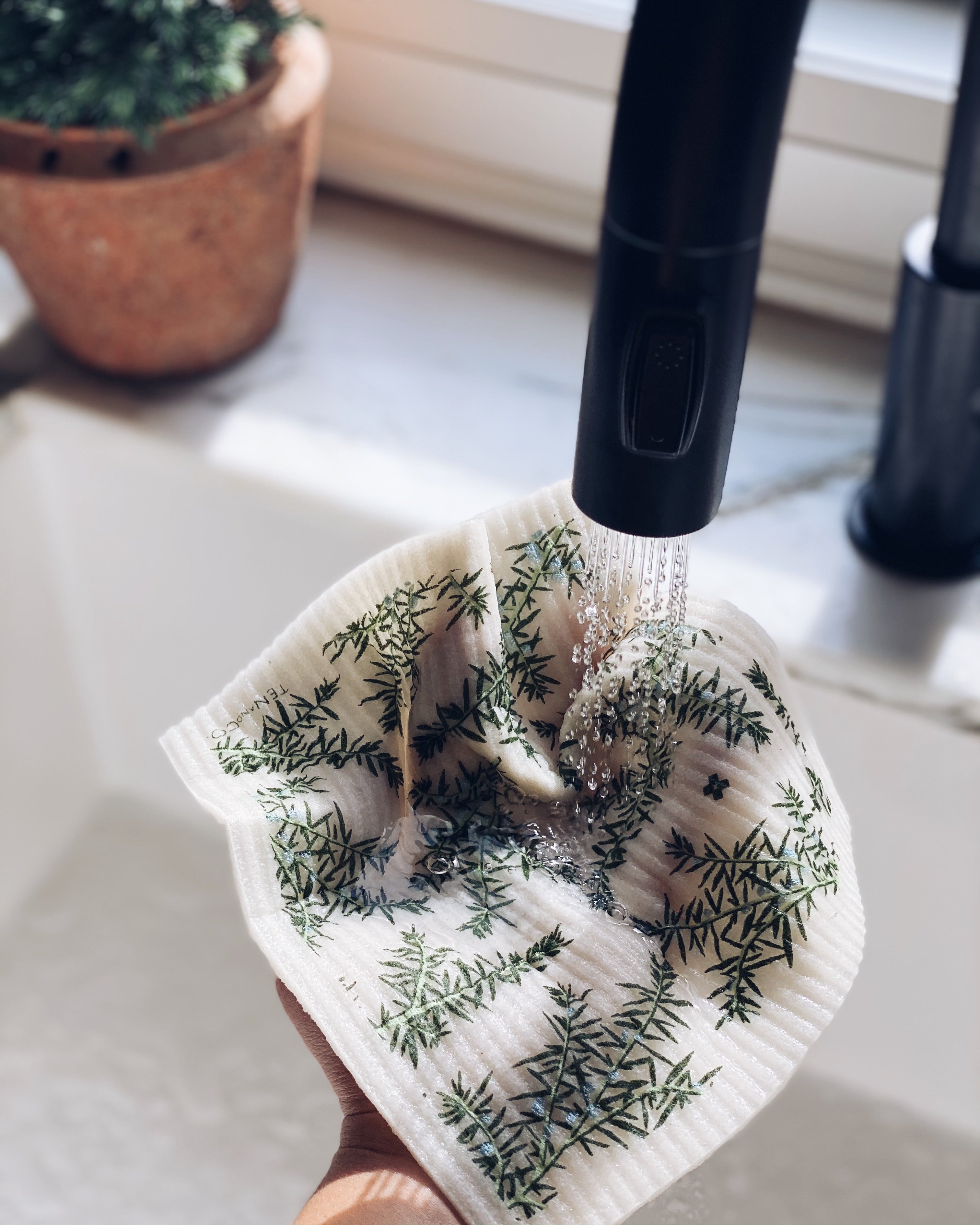 Product image of Juniper Greens on White sponge cloth. There is a white kitchen sink with a black faucet. There is a hand holding the Juniper Greens on White sponge cloth under the running water. There is a small plant on the edge of the sink.