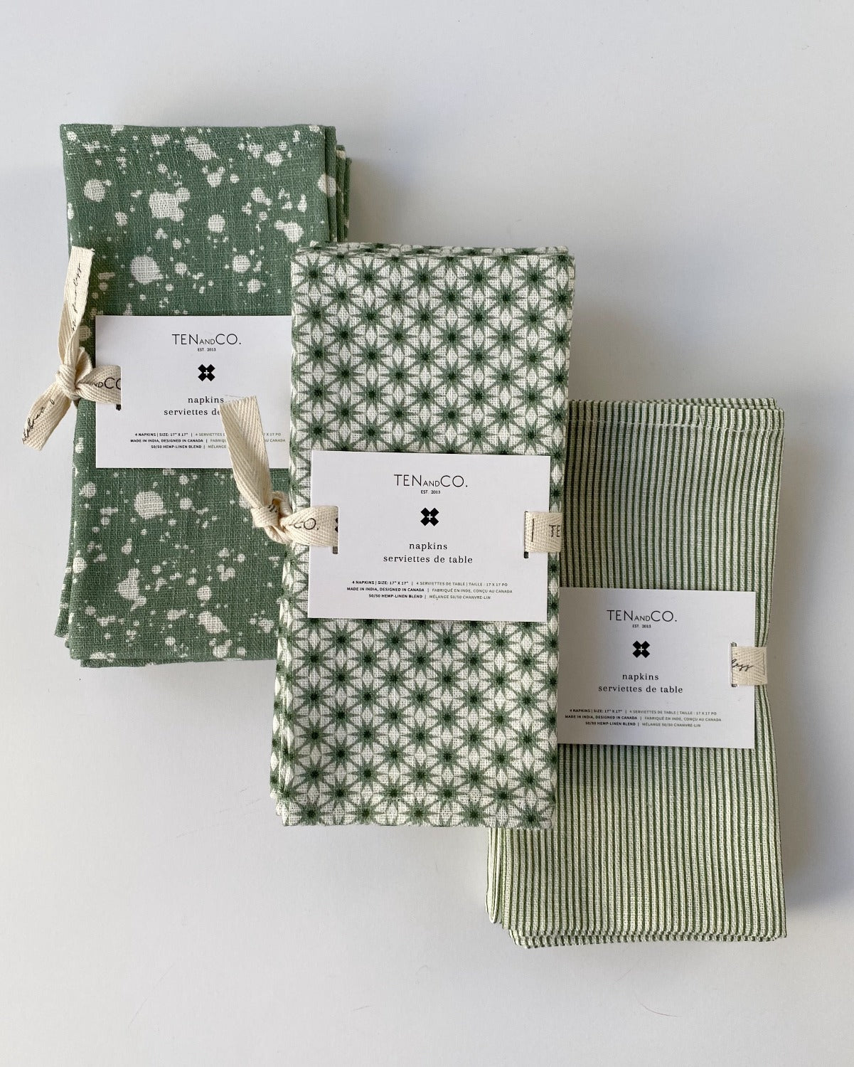 Flat lay image of 3 different sets of napkins. From left to right; Splatter Sage, Starburst Sage, Stripe Sage. All sets of napkins are laid in a diagonal method, with the Splatter Sage closer to the top left, and the Stripe Sage closer to the bottom right of the image. All napkins are tied with the Ten and Co printed description label on top.