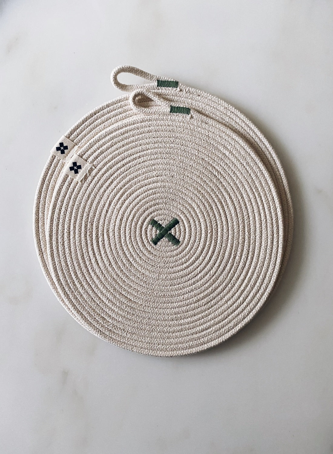 Flat lay image of Large Rope Trivet Sage. The white rope trivet is laying on a white marble surface. The trivet is a large spiral with a sage/green X in the middle. The smaller trivet is on top of it. There are tiny Ten and Co. Logo attached to each side. There is a loop at the end of the rope to use for hanging.    