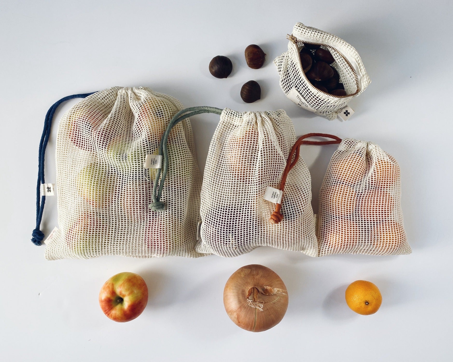 Flay lay image of Produce Bags on a white background. In this image there the 3 different sized produce bags each filled with different produce. There are apples, onions and clementines. There is also a small pouch that has figs in it and there are 3 sitting beside it.