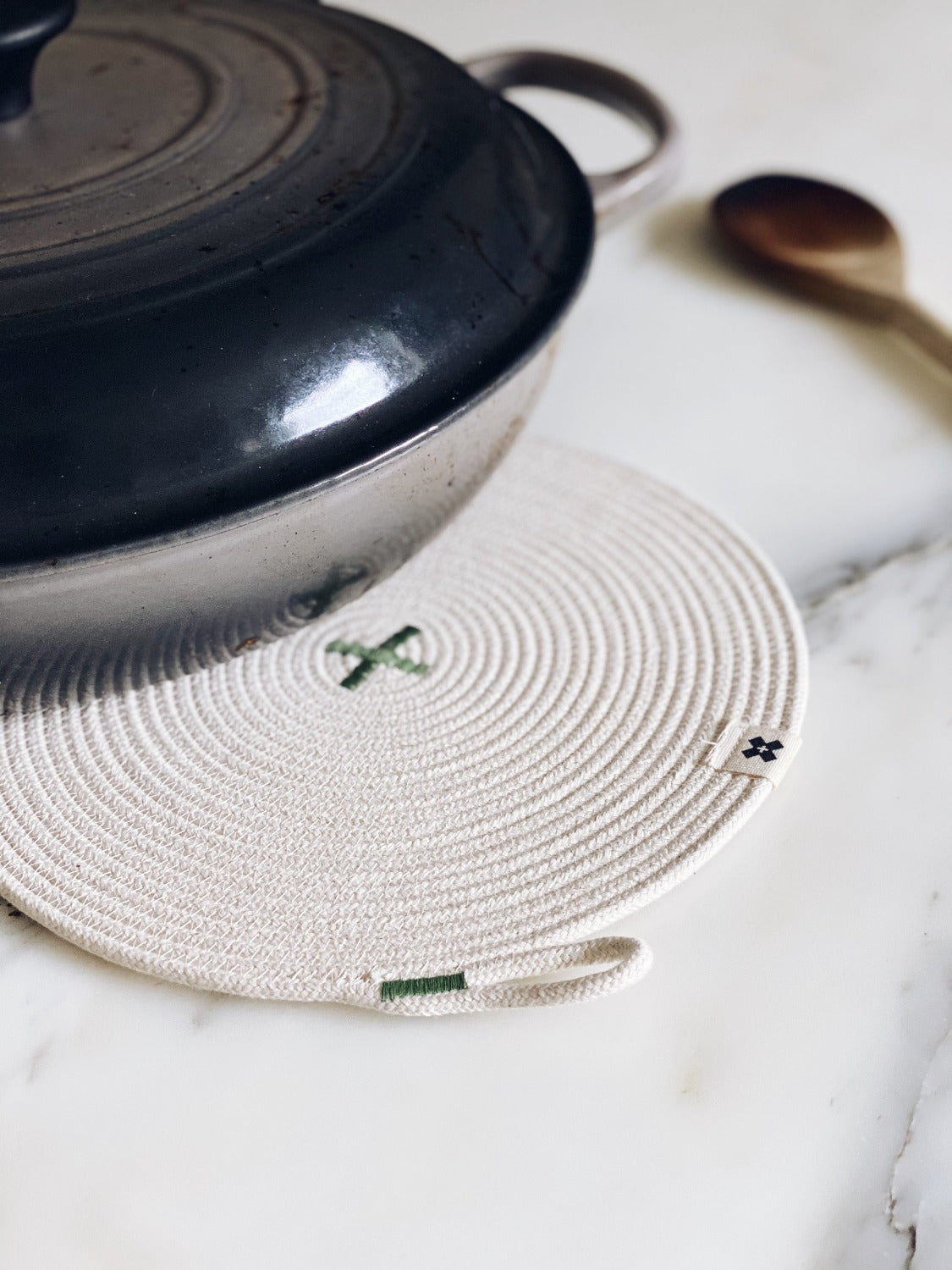 Flat lay image of Large Rope Trivet Sage. The white rope trivet is laying on a white marble surface. There is a black pan with a black lid on the edge of the trivet. There is a wooden spoon lying next to the trivet and pan. 