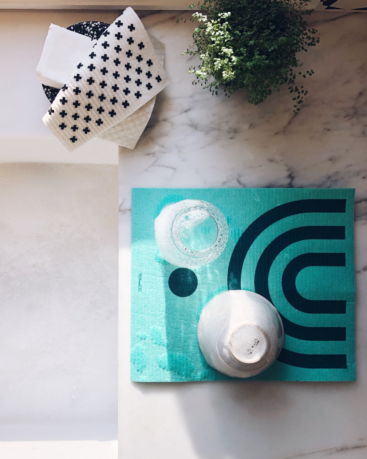 Aerial product image of LARGE Arc Green Sponge Cloth Mat. There is a marble counter beside a white kitchen sink. On the counter is a LARGE Arc Green sponge cloth mat with a glass and a bowl upside down, drying. There is a soap dish with a Tiny X sponge cloth on it. Beside the soap dish is a small green plant. There is sun shining on the sponge cloth mat. 