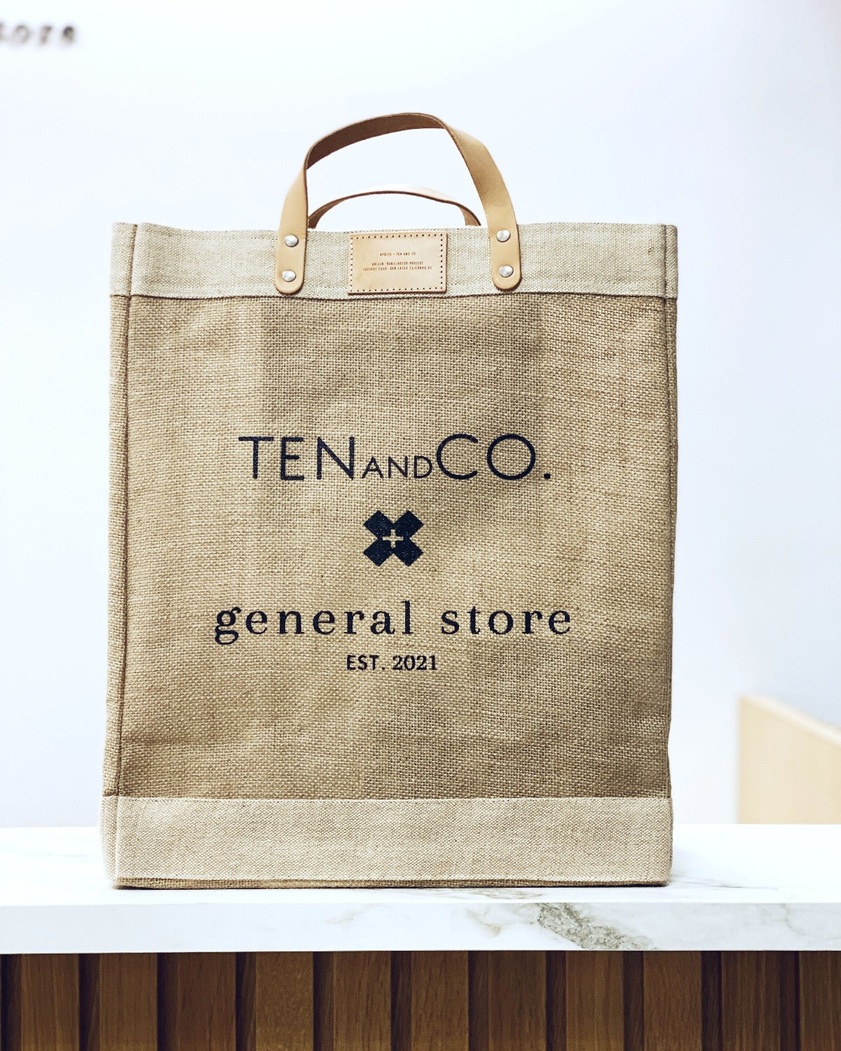 Flat lay image of Market Bag – Natural on a white background. This market bag is made of golden jute fiber with leather straps . The front says Ten and Co. General store in black font with an X in the middle. There is a leather tag at the top by the straps. 