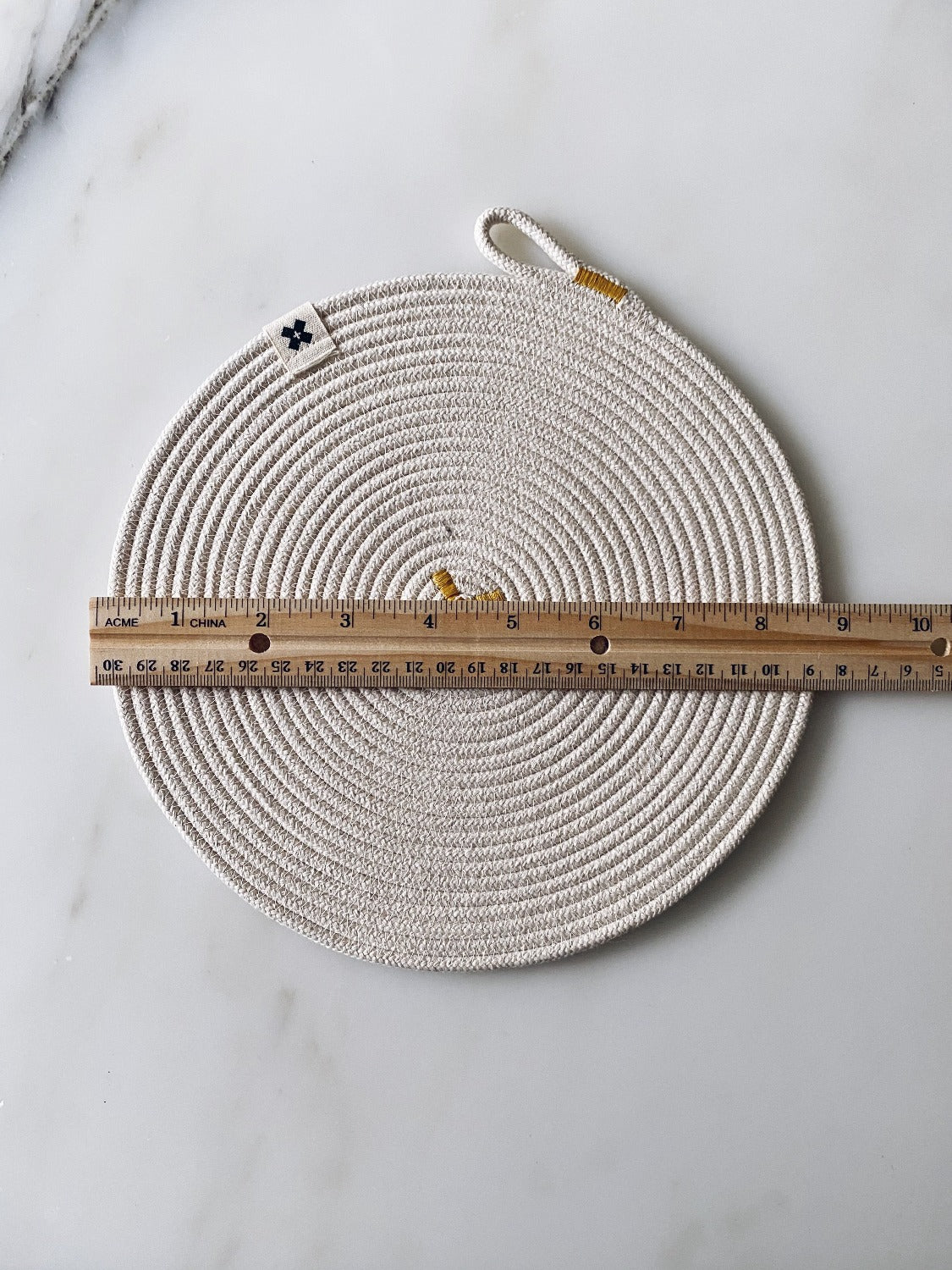 Aerial image of Large Rope Trivet Ochre. On a marble surface there is an Ochre trivet. There is a wooden ruler across the trivet. It is around 9 inches in diameter. 