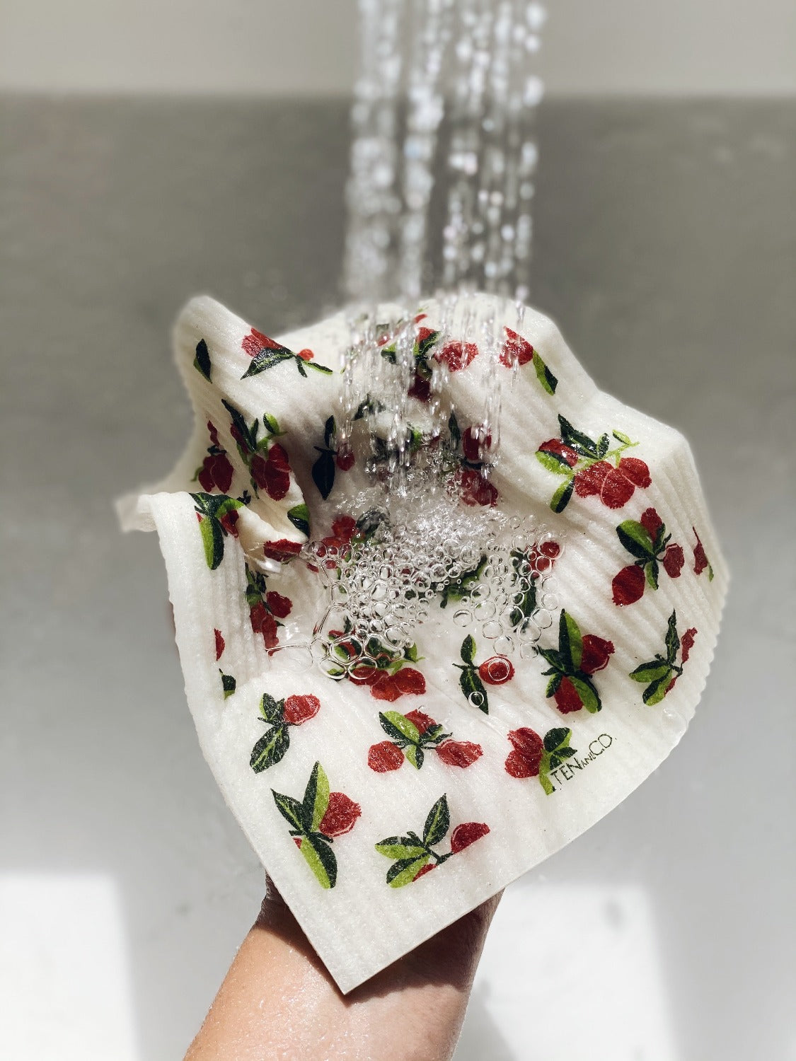 Design image of Cranberry Sponge Cloth. Image is zoomed on the water stream from a sink coming from the top of the image. Sink is white and cloth is being held from underneath, hand coming from the bottom of the image. Water is pooling on top of the cloth.
