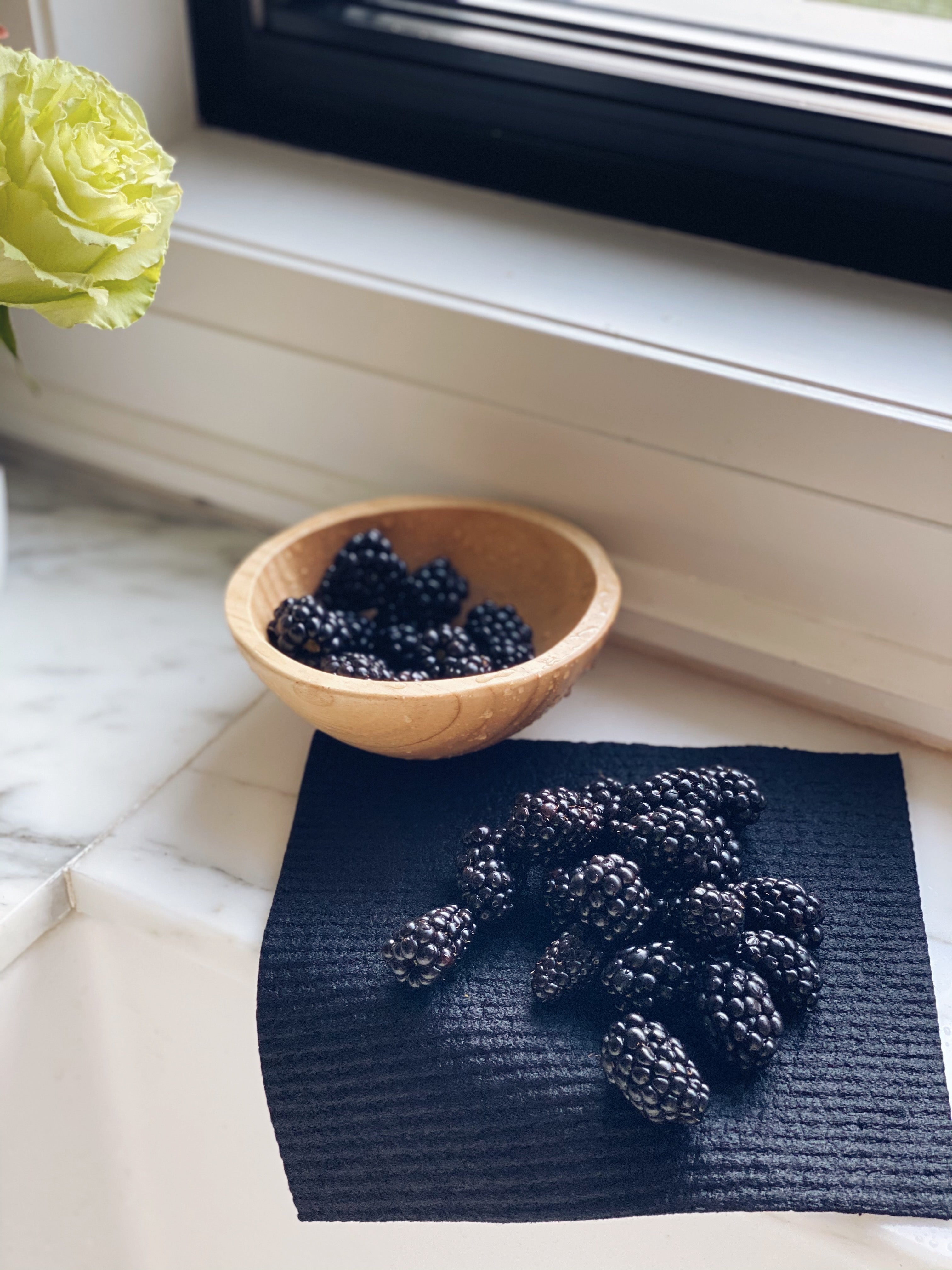 Solid Black Sponge Cloth hangs over the back of the sink.  On top of the cloth are freshly washed Blackberry that are drying on the cloth.  To the top left of the image is a wooden bowl  of blackberries.  In the background you can see the light streaming onto the window sill and on the far left is an image of green roses cut off.