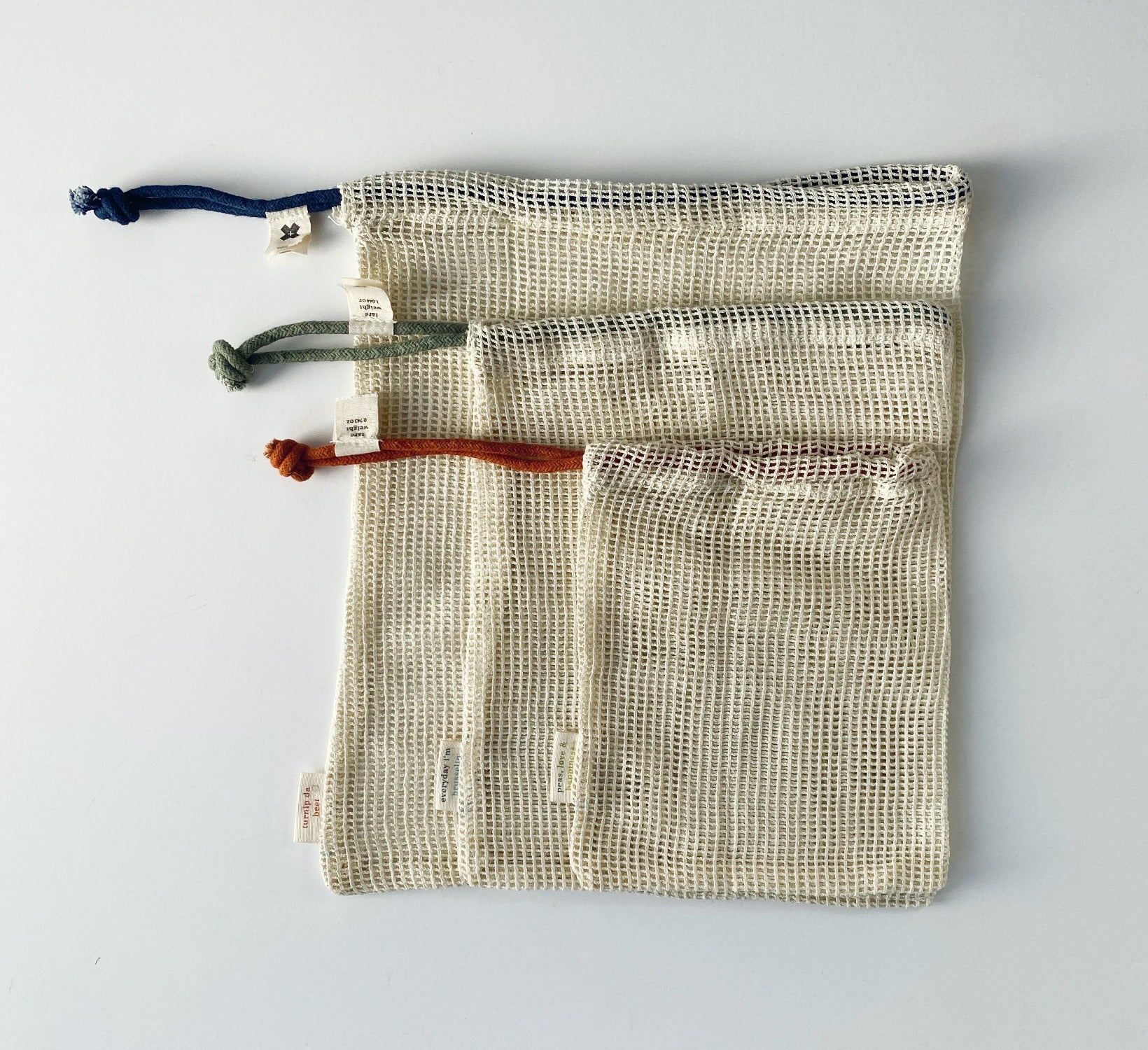 Flat lay image of Produce Bags on a white background. In this image there are 3 produce bags in 3 different sizes. Small, medium and large. The small bag has a rust colour string, the medium has a sage colour string and the large has a denim colour string. They are laying on top of each other to show the difference in sizes They are cotton mesh. They also have small tags with the Ten and Co. Logo on them. 