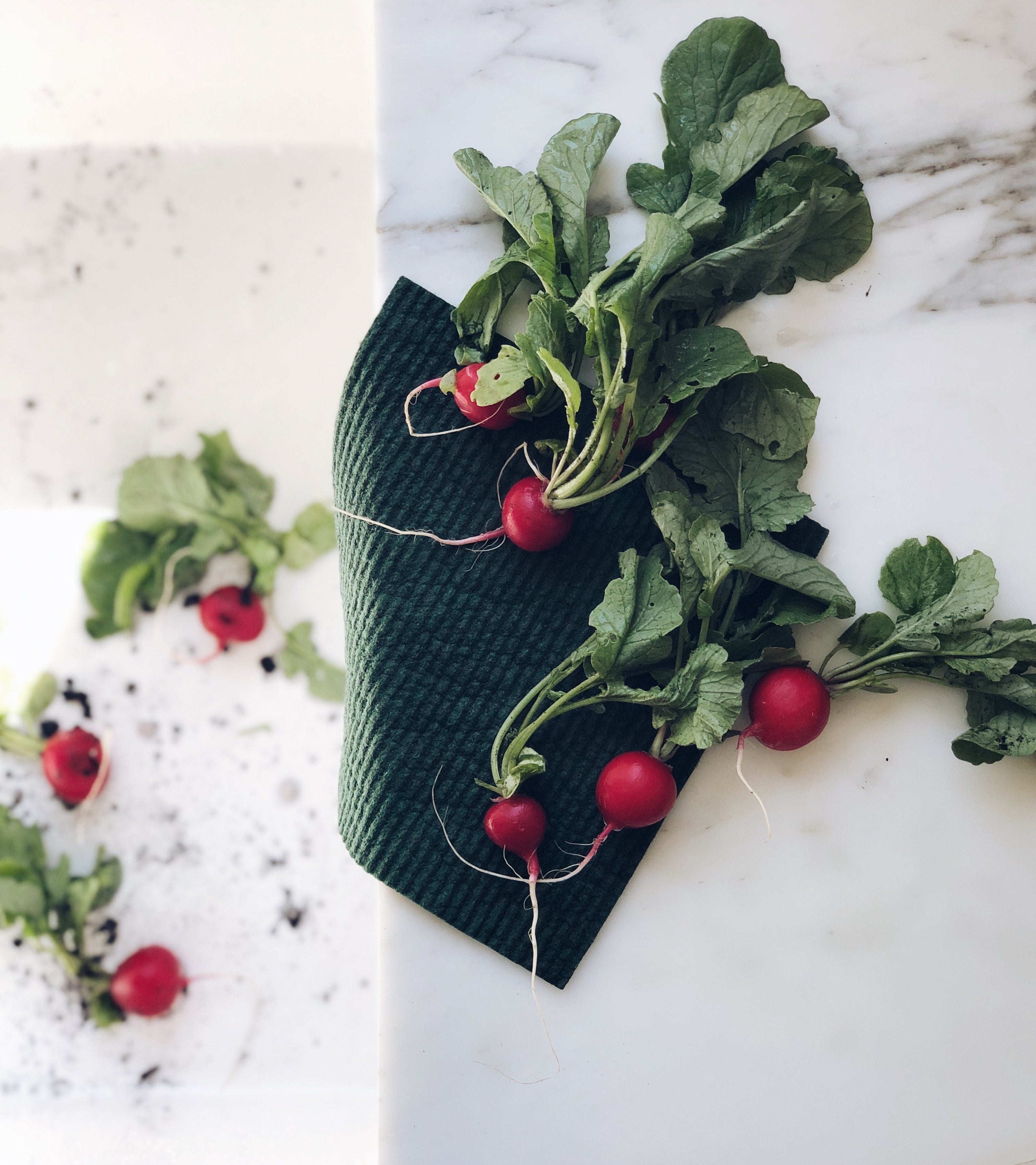 Radishes lying on top of a damp solid evergreen sponge cloth.  In the sink you can see radishes that still have dirt on them and need to be cleaned. This is on top of a marble counter top beside a sink.