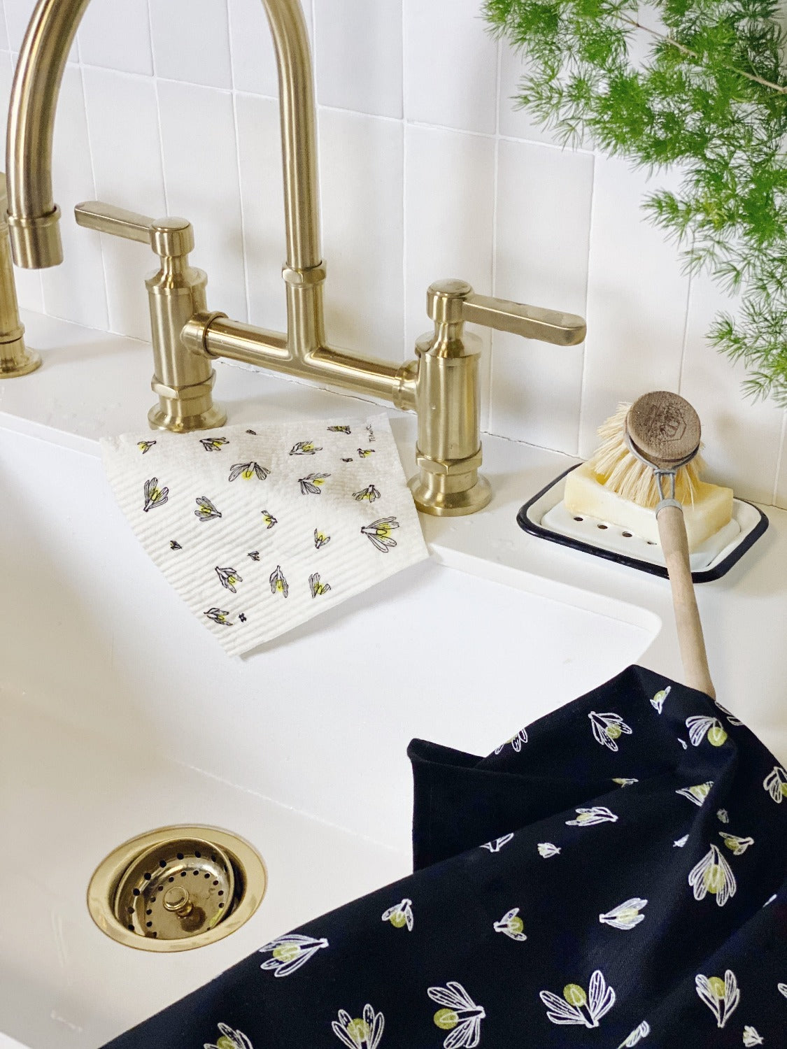Product image of Firefly gift set on a black background. In this image there is a white kitchen sink with brass fixtures. There is a Firefly sponge cloth laying over the edge of the sink near the faucet. Beside the faucet is an enamel soap dish with a bar of soap and a dish brush. On the side edge is the black firefly tea towel. There is a small green plant in the top right corner.  
