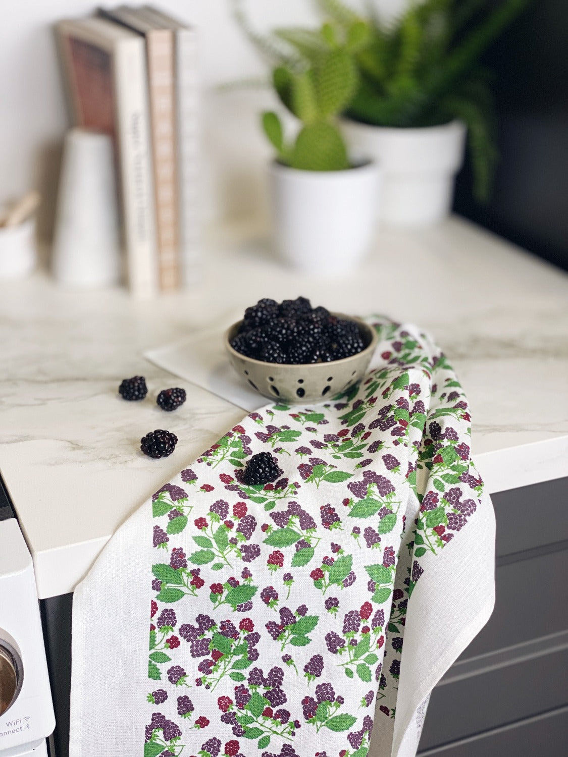 Product image of Vintage Blackberry gift set. In this image there is a white marble countertop with grey cabinets underneath. Against the back, out of focus, are 3 books held up with book ends and two small green plants in white pots. There is a vintage blackberry tea towel with a small bowl full of blackberries on it. There are a few blackberries sprinkled on the counter as well. You can see a little bit of a stove on the left side. 
