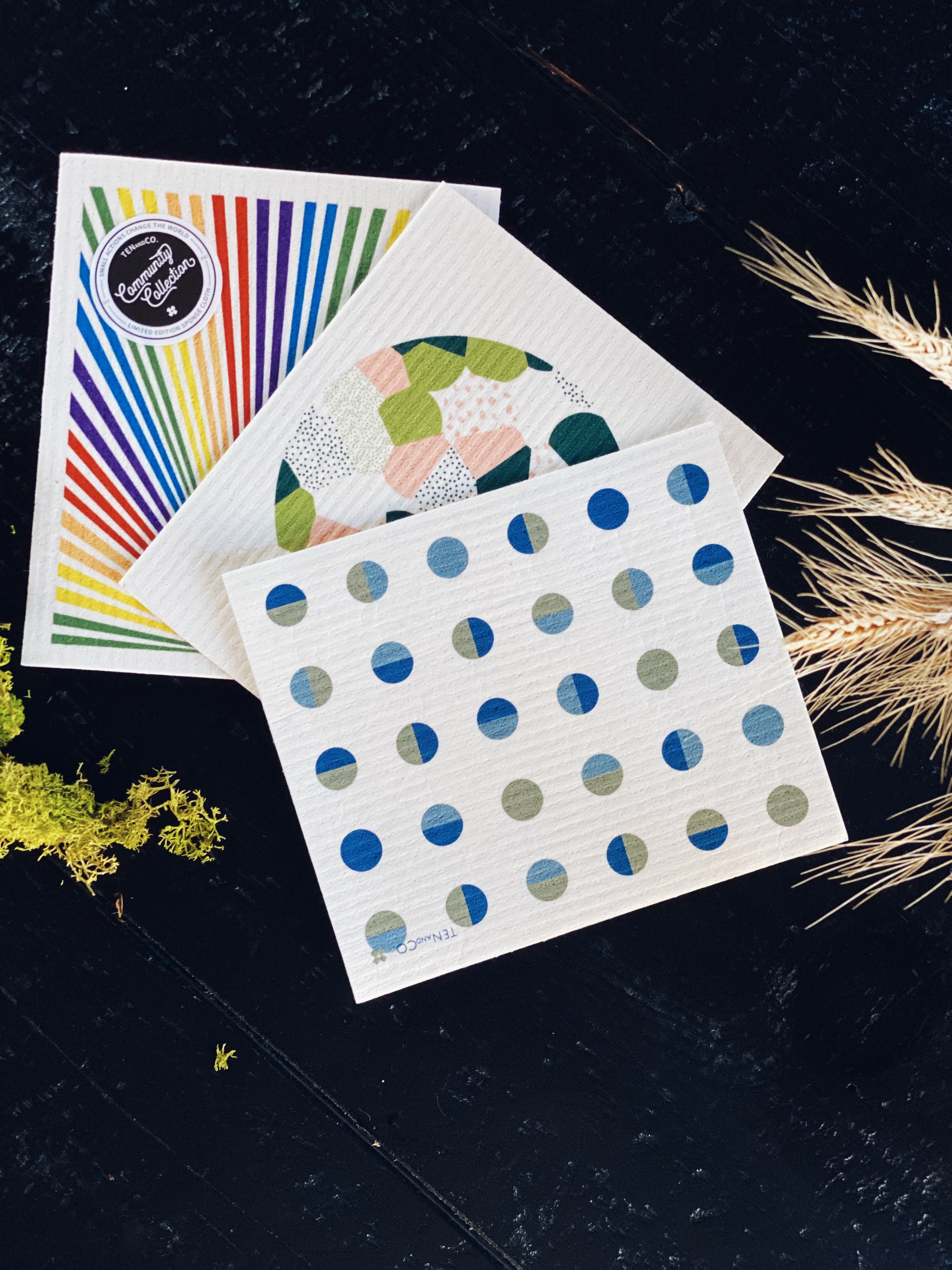 Aerial product image of Moon Sponge Cloth on a black background. In this image there are 3 different sponge cloths from The Community Collection. From left to right: Pride, Full Circle and Moon. There is some green moss and white straw feathers. 