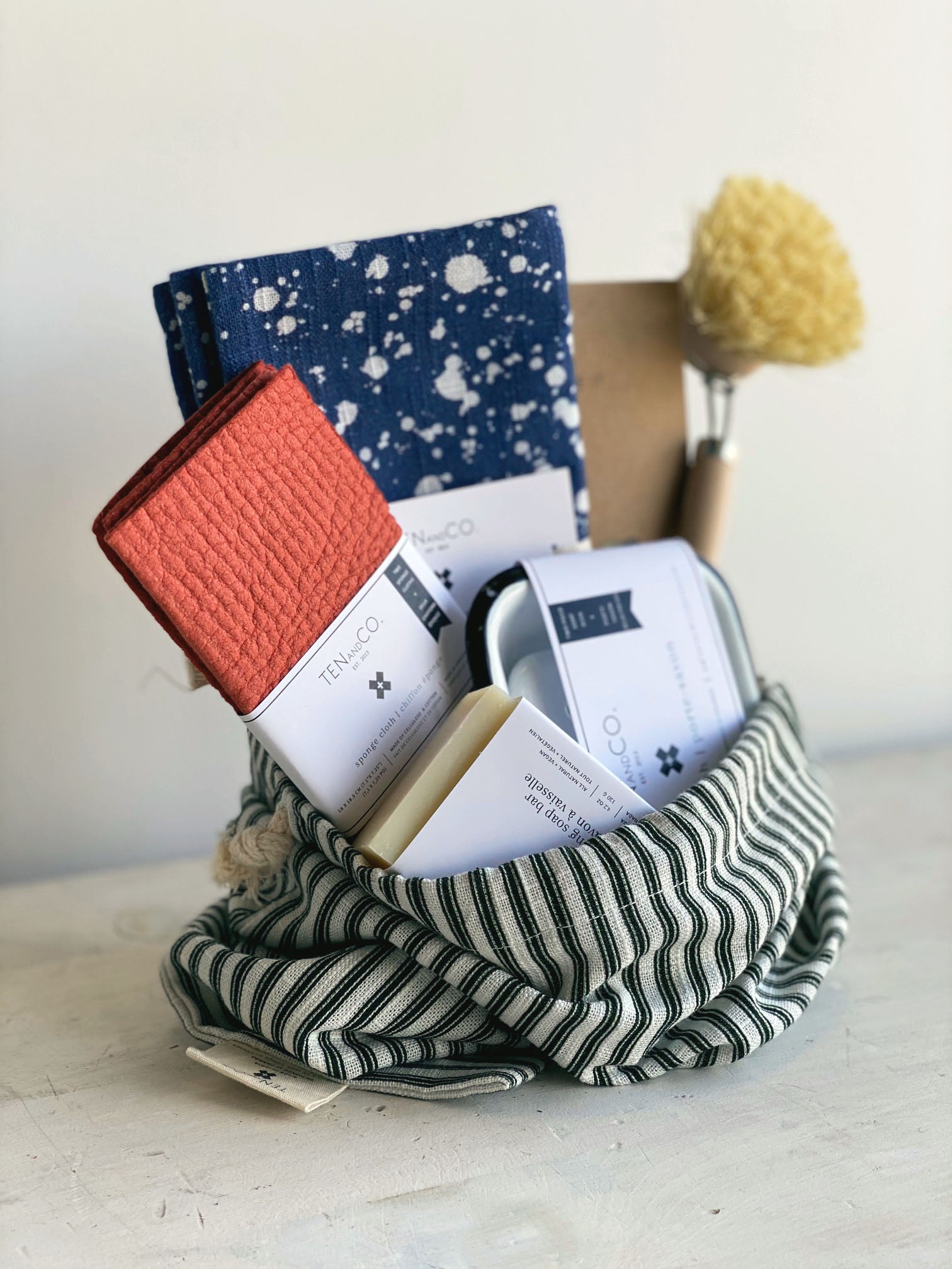 Angled photo of Gift Bag with Ten and Co items inside. Photo on a white surface against a white wall. Deep green and white vertical striped gift bag is propped open with the following items inside. Left to right; Rust solid 2pk, Splatter Denim Napkins, Beeswax Wraps, Scrub Brush. In front of these items in the bag, is a Dishwashing Soap Bar and an Enamel Soap Dish. 