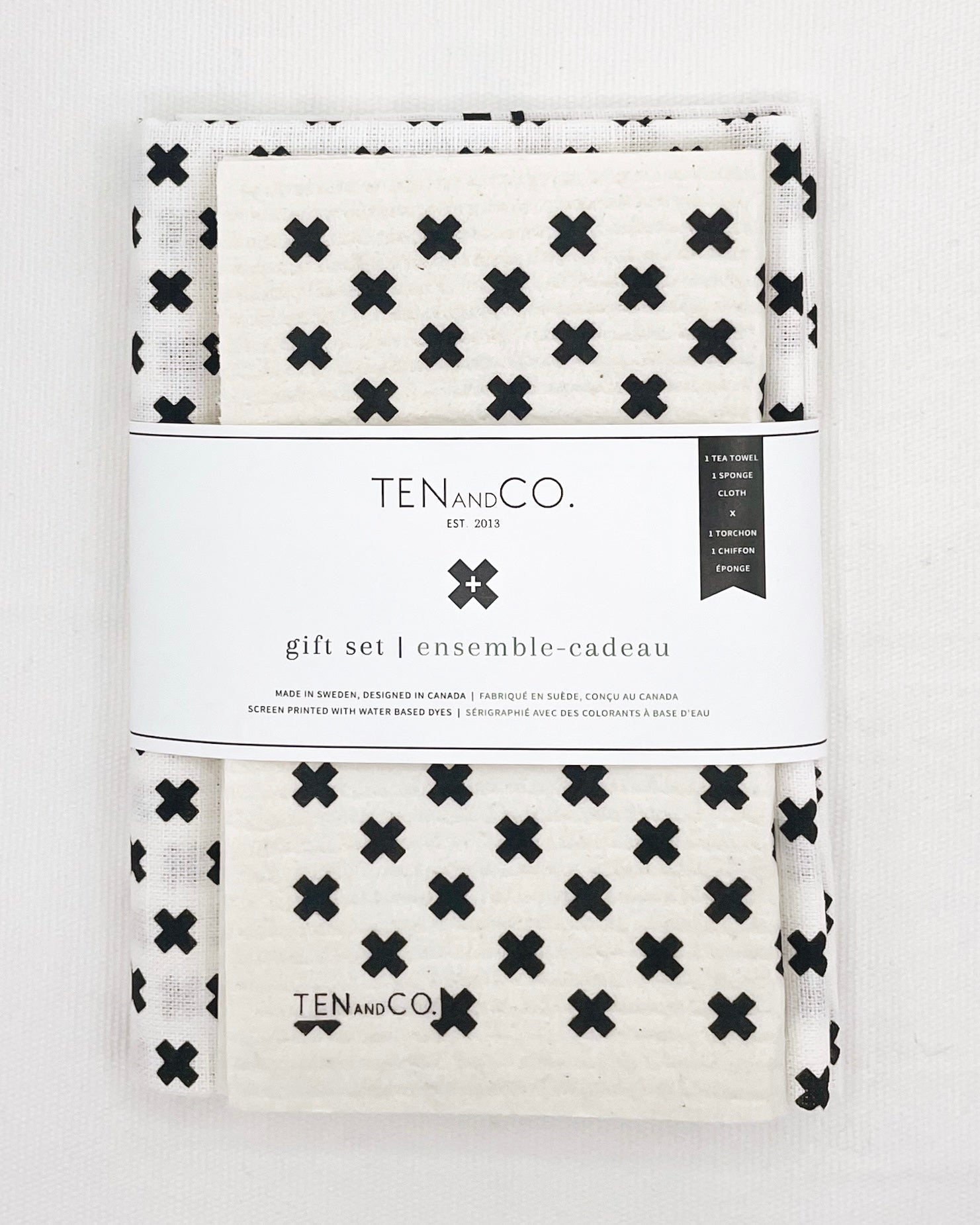 Flat lay image of Tiny X Black on White gift set on a white background. There is a Tiny X Black on White sponge cloth folded with a Tiny X Black on White tea towel behind it. There is a white belly band across both. The sponge cloth has a white base with tiny x’s in black throughout. The tea towel has a white base with tiny x’s throughout. The belly band has the Ten and Co. Logo and descriptive text in black font.