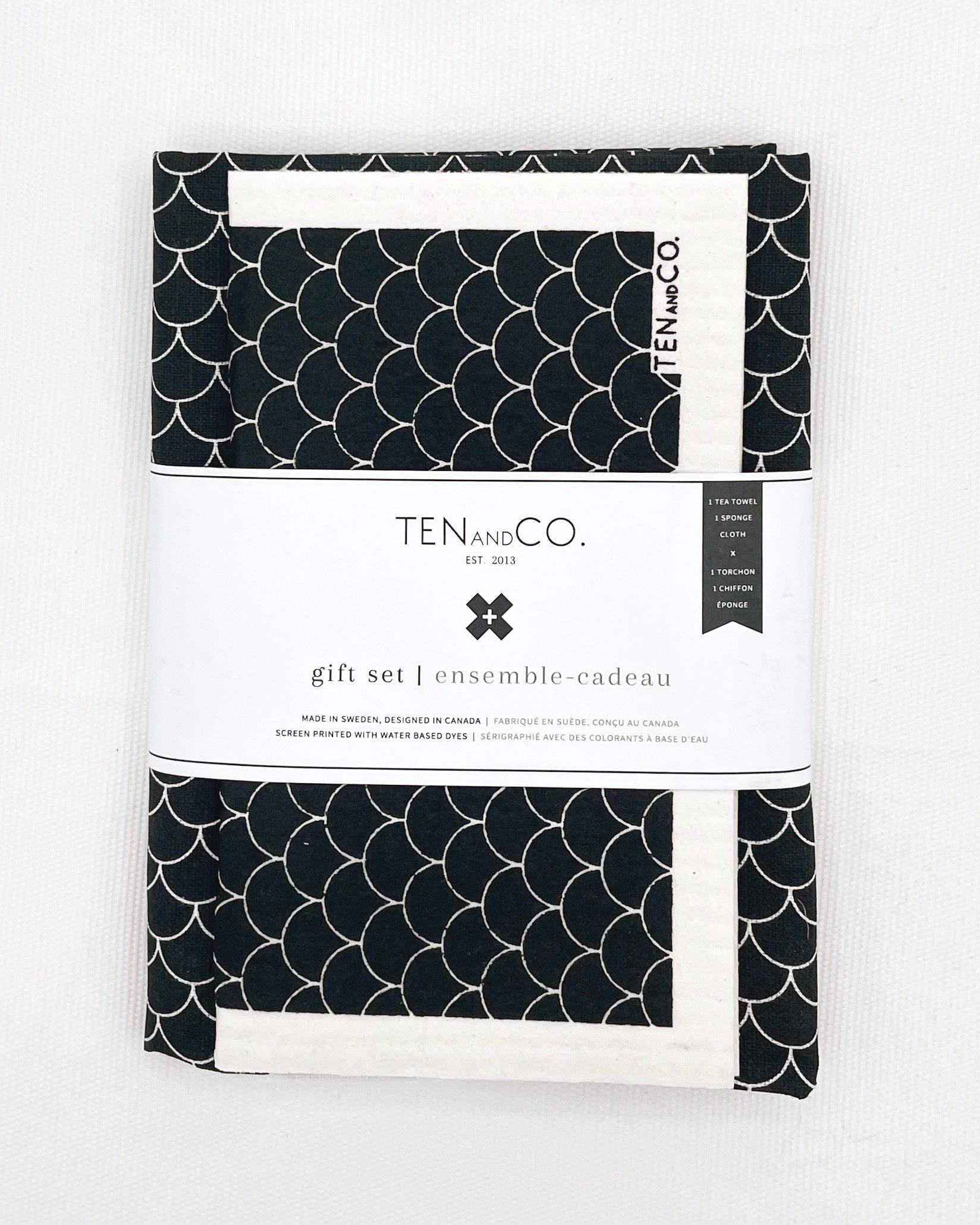 Flat lay image of Scallop Black gift set on a white background. There is a Scallop Black sponge cloth folded with a Scallop Black tea towel behind it. There is a white belly band across both. The sponge cloth has a white base with a black background that has white scallops throughout. The tea towel has a white base with black background and white scallops throughout. The belly band has the Ten and Co. Logo and descriptive text in black font.
