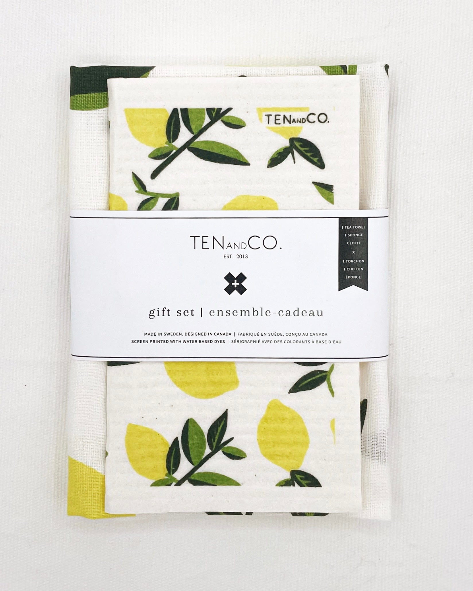 Flat lay image of Citrus Lemon Gift Set on white background. There is a Citrus Lemon sponge cloth folded with a Citrus Lemon tea towel folded behind it. They have a white belly band around them. The sponge cloth has yellow lemons with a green stem throughout. The belly band has the Ten and Co. Logo with descriptive text in black font.