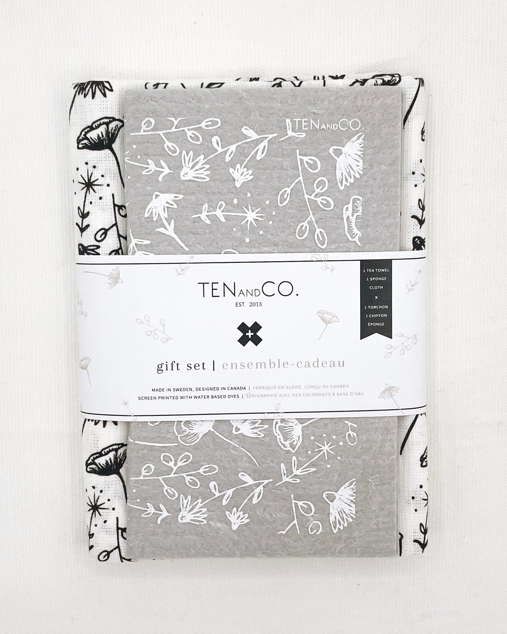 Flat lay image of Modern Floral gift set. There is a Modern Floral sponge cloth folded with a Modern Floral tea towel behind it. There is a white belly band across both. The sponge cloth has a grey base with white flowers throughout. The tea towel has a white base with black flowers throughout. The belly band has the Ten and Co. Logo and descriptive text in black and grey font.